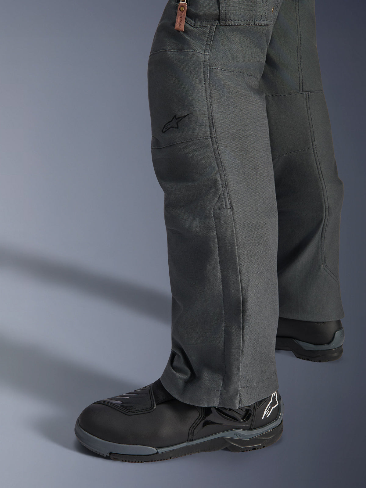 Pantalon Alpinestars Borrego Drystar®, pantalon de moto d'aventure gris anthracite avec un aspect toile cirée, construction textile vue de profil avec des bottes de moto noires, conception robuste pour la route et le touring d'aventure