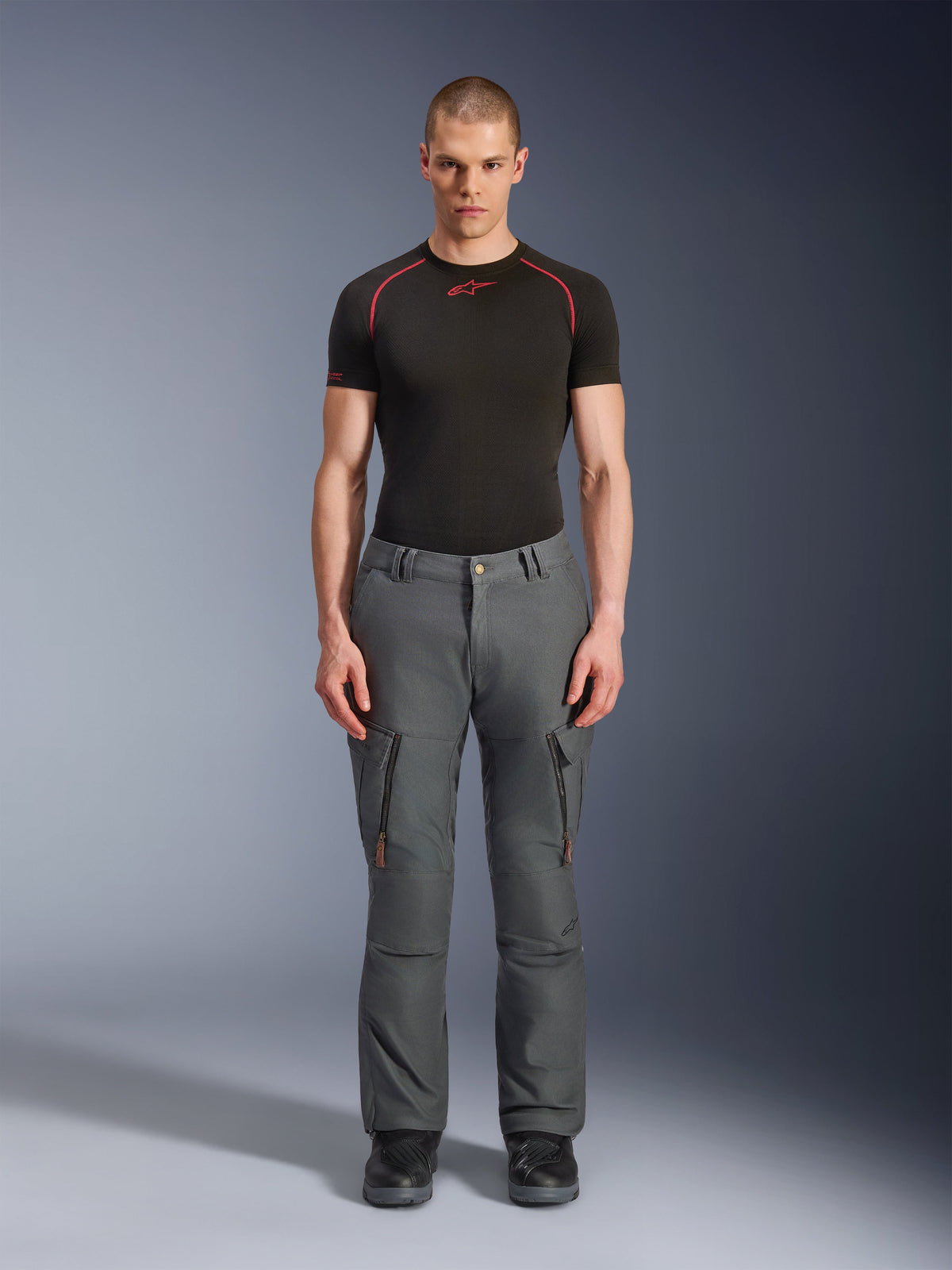 Pantalon Alpinestars Borrego Drystar®, pantalon de moto d'aventure gris, porté par un mannequin avec un t-shirt technique noir et rouge et des bottes noires, doté de poches cargo avec fermetures éclair verticales, aspect toile cirée robuste, conception durable pour le tourisme d'aventure et la conduite sur route