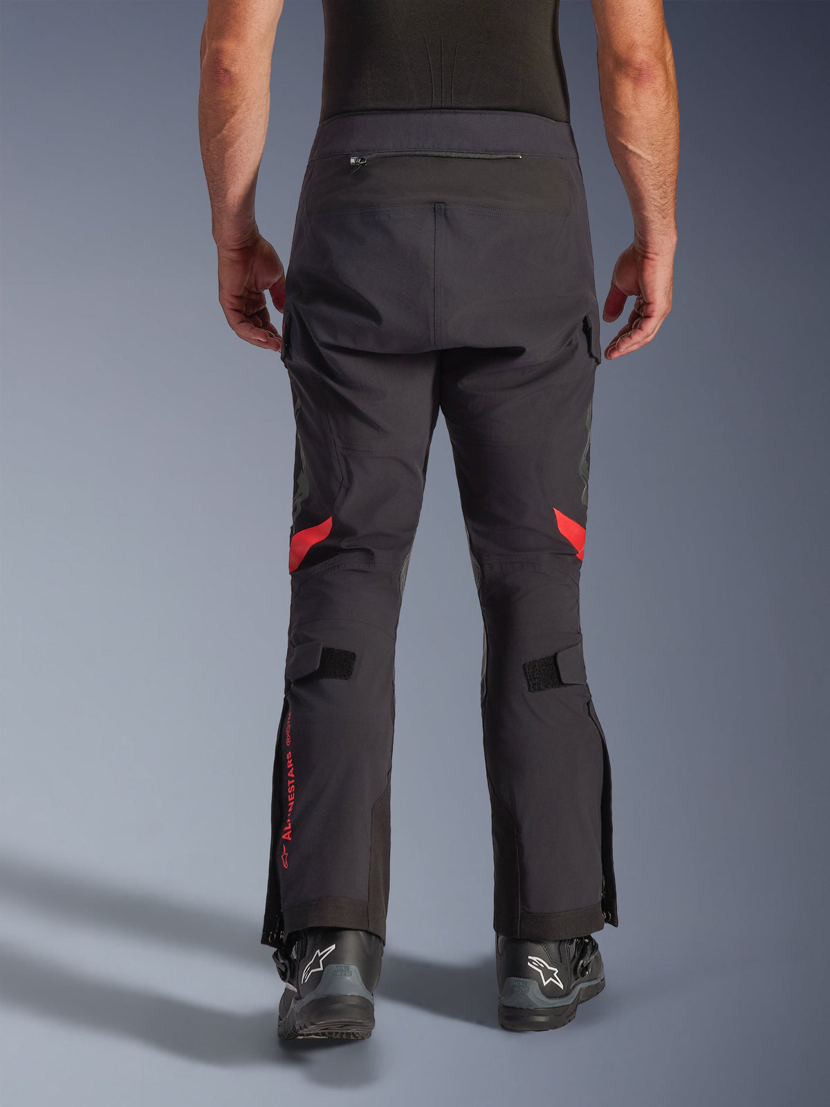 Alpinestars Halo Pro Drystar® XF Laminierte Hose, Black Dark Shadow, Rückansicht einer Adventure-Motorradhose mit roten Akzenten in den Kniekehlen und rotem Branding an der Wade, kombiniert mit schwarzen Alpinestars-Stiefeln, strapazierfähiges 3L-laminiertes Textil für All-Terrain-Performance