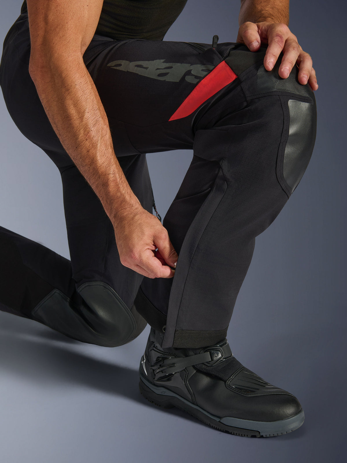 Alpinestars Halo Pro Drystar® XF Laminierte Hose, Black Dark Shadow, Nahaufnahme des Unterschenkels mit verstellbarem Waden-Reißverschluss, strapazierfähige Knieverstärkungen, rote Akzente, 'astars'-Branding, getragen mit schwarzen Motorradstiefeln