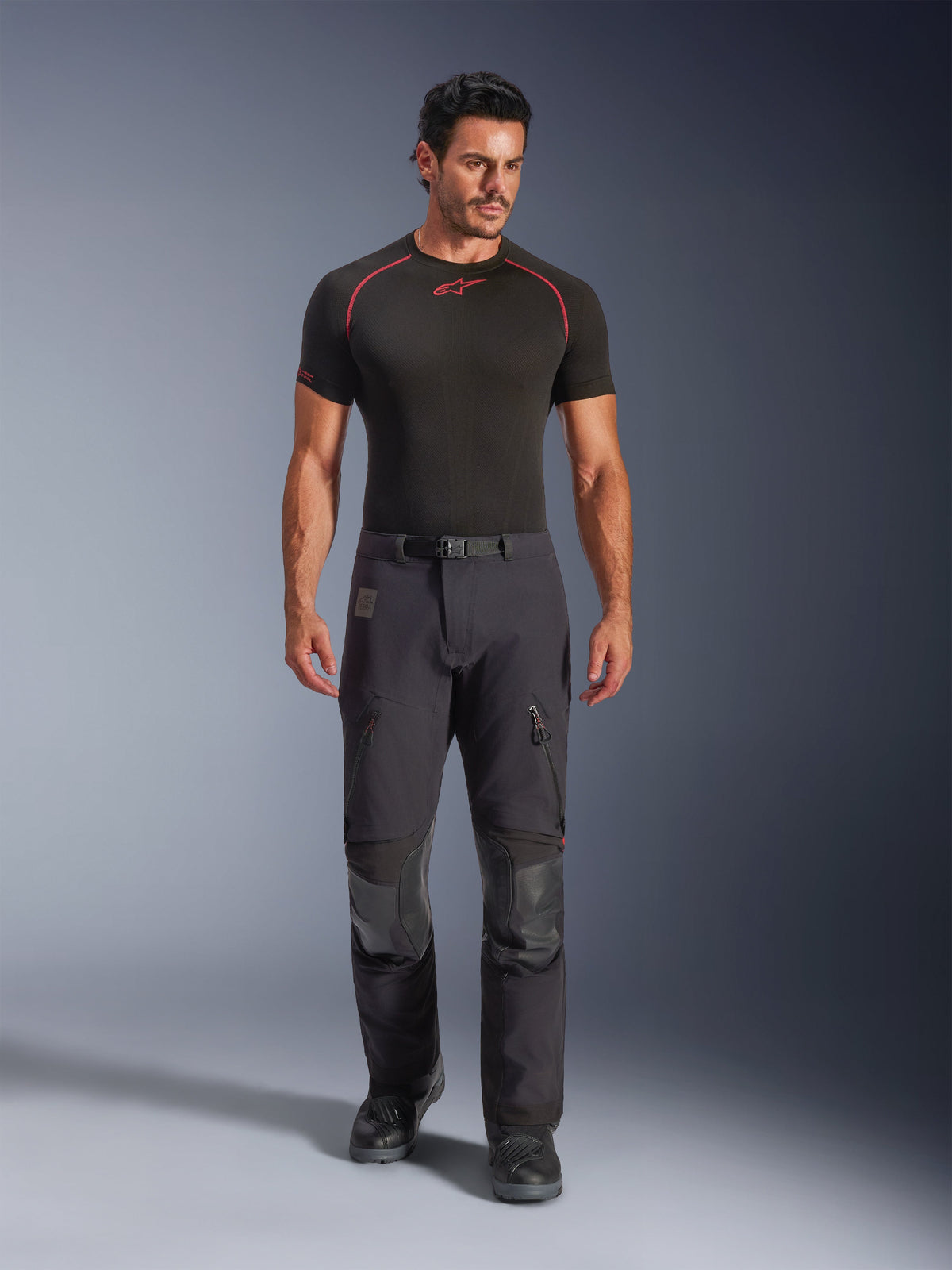 Alpinestars Halo Pro Drystar® XF Laminierte Hose, Black Dark Shadow, getragen von einem Model mit schwarzem Base-Layer und Motorradstiefeln, mit technischen Belüftungsreißverschlüssen am Oberschenkel, Cargotaschen und verstärkten Knieeinsätzen, Adventure-Touring-Motorradausrüstung.