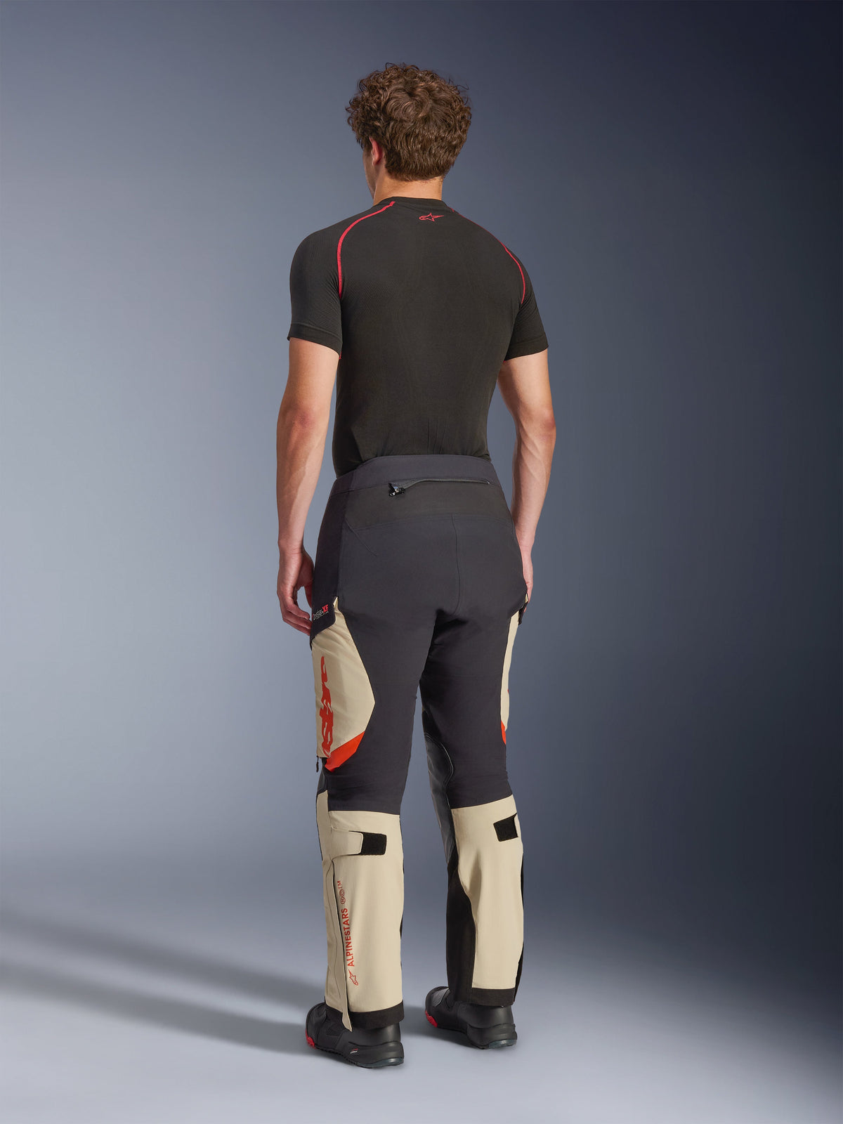 Alpinestars Halo Pro Drystar® XF Laminierte Hose, Adventure-Motorradhose, Mastic Black und Tan mit orangefarbenen Akzenten, Rückansicht zeigt die 3-lagige laminierte Schalenkonstruktion, kombiniert mit einem schwarzen Base-Layer und Adventure-Stiefeln.