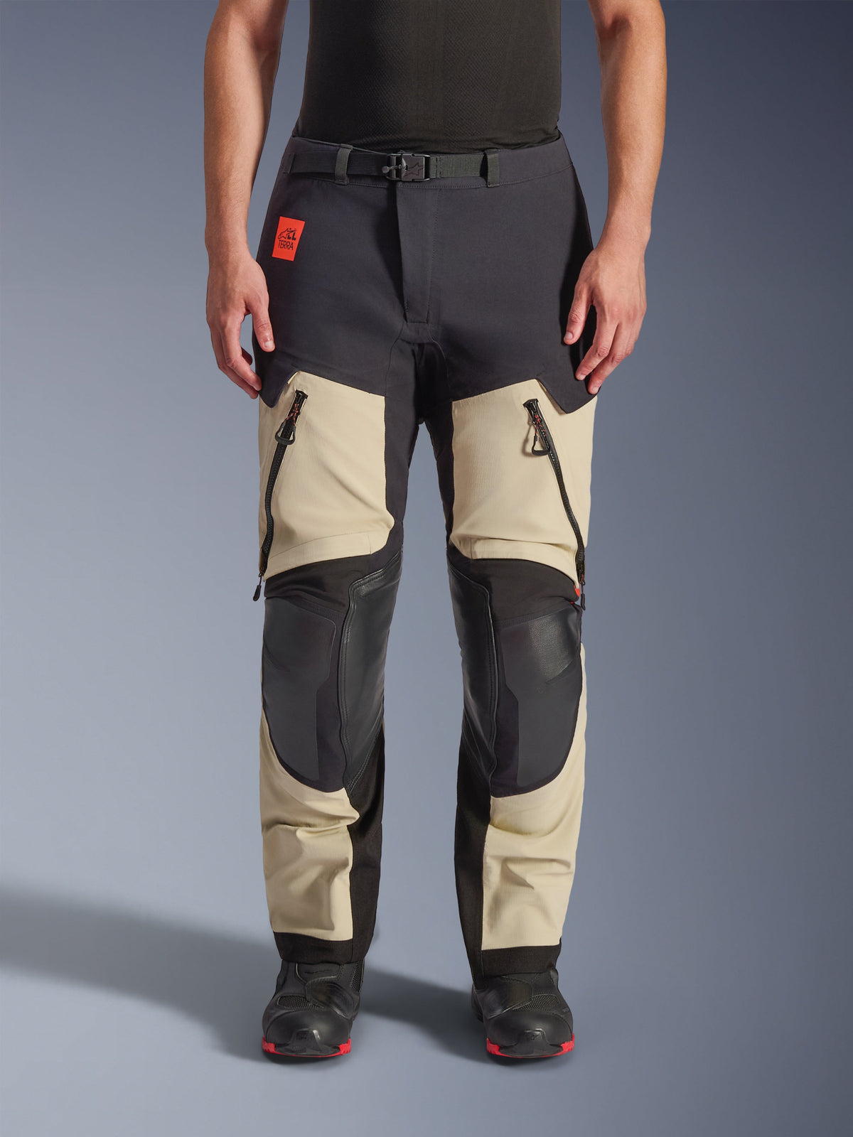 Alpinestars Halo Pro Drystar® XF Laminierte Hose, Adventure-Motorradhose, Mastic Black, Vorderansicht getragen, mit großen Belüftungsöffnungen mit Reißverschluss an den Oberschenkeln, verstärkten schwarzen Kniepaneelen und sandfarbener Textilkonstruktion, kombiniert mit schwarz-roten Motorradstiefeln