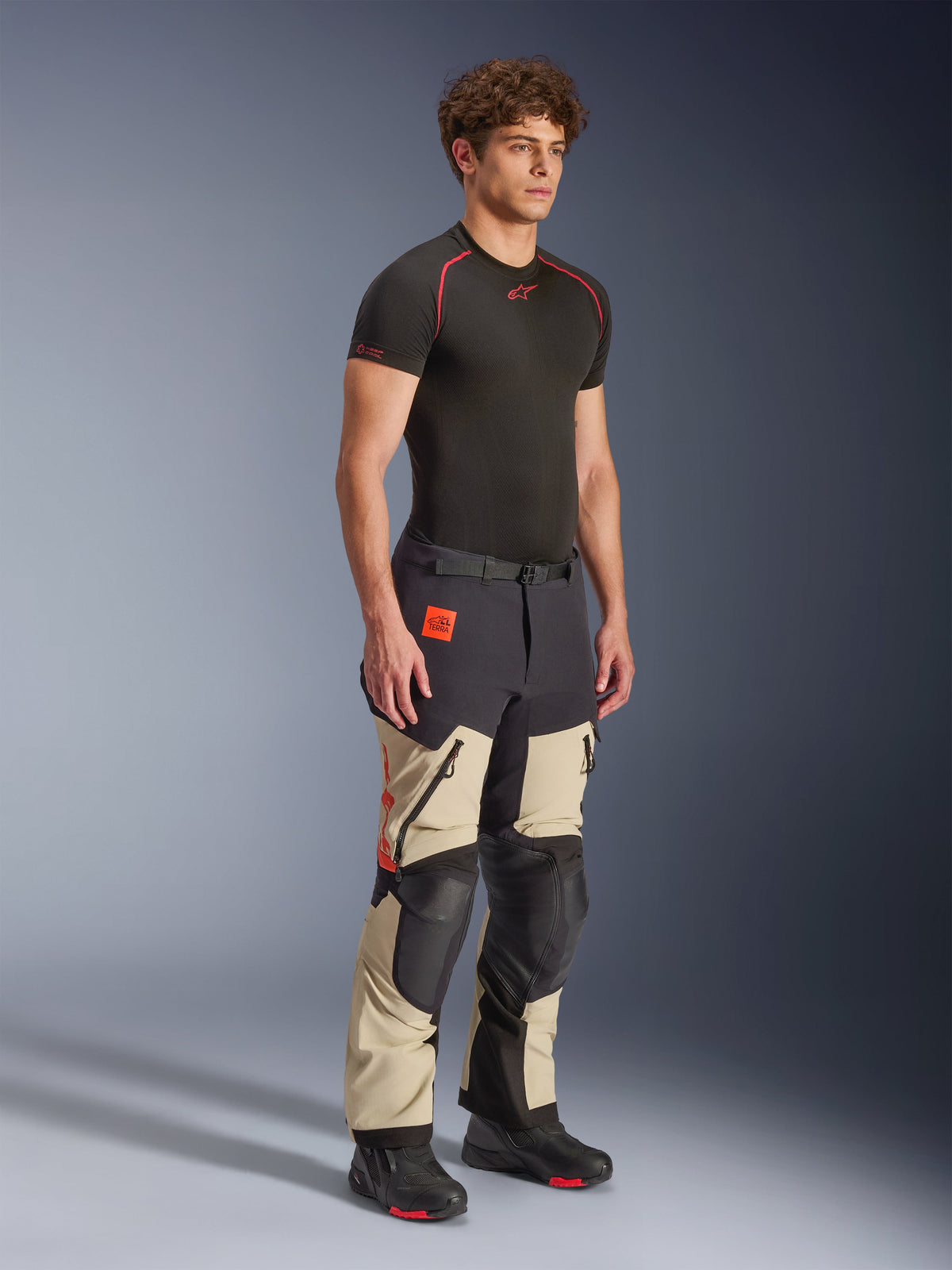 Alpinestars Halo Pro Drystar® XF Laminierte Hose in Mastic Black, Adventure-Motorradhose, getragen von einem Modell, mit schwarzen Knie- und Gesäßverstärkungen, orangefarbenem Branding und mehreren funktionellen Reißverschlüssen, kombiniert mit einem schwarzen Base Layer und Motorradstiefeln.