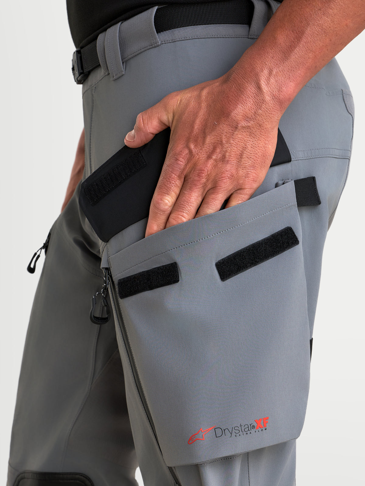 Alpinestars Maxdura Drystar XF Laminated broek, adventure motorbroek in donkergrijs en zwart, met detail van een hand die de schuine dijbeenzak opent met Drystar XF branding