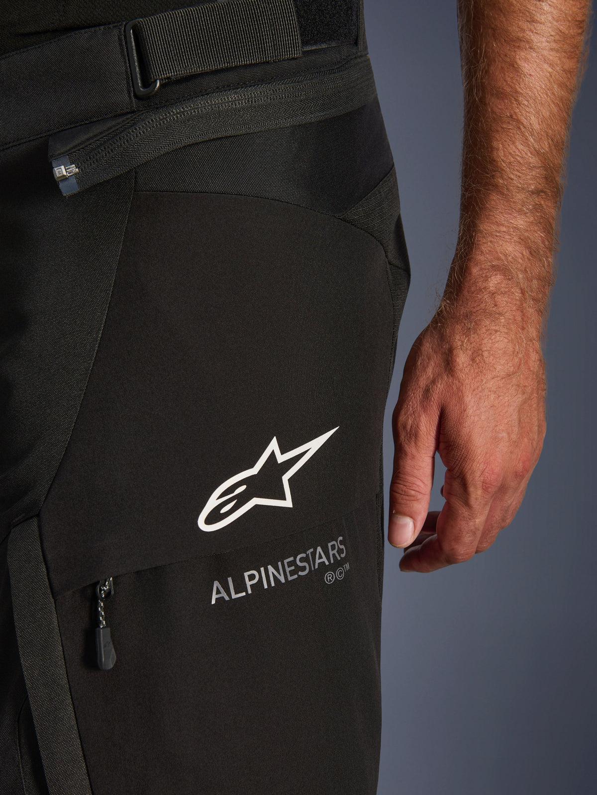 Détail en gros plan du pantalon Alpinestars Andes V4 Drystar® noir, présentant le logo et le lettrage Alpinestars blancs sur le rabat d'une poche cargo, avec sangle de réglage de la taille et fermeture éclair de connexion visibles, conçu pour le touring d'aventure à moto