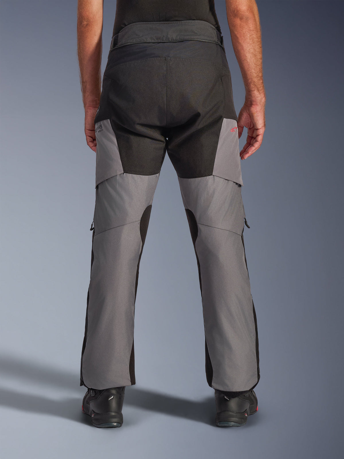 Pantalon Alpinestars Andes V4 Drystar®, pantalon de moto aventure, noir et gris foncé, vue arrière montrant l'assise noire renforcée et les jambes grises, avec un petit logo rouge, associé à des bottes de moto noires, construction textile durable pour le touring par tous les temps.