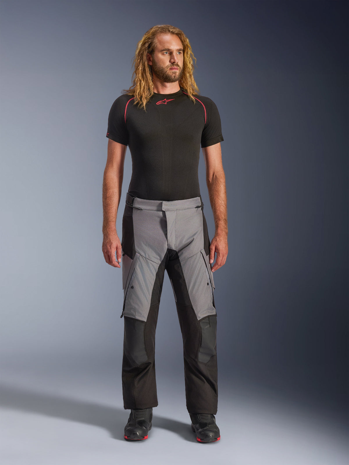 Alpinestars Andes V4 Drystar® broek, zwart en donkergrijs, adventure motorbroek gedragen door een model met een zwarte basislaag en zwarte laarzen, voorzien van een textielontwerp met meerdere panelen met grijze voorvlakken en zwarte verstevigingen, ontworpen voor toertochten in alle weersomstandigheden en rijden op de weg.