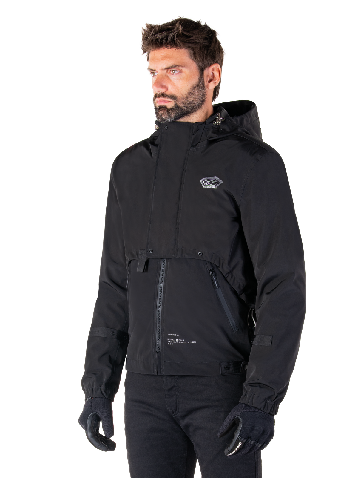 Alpinestars Mootant Waterproof Parka, schwarze modulare Motorradjacke, getragen von einem Mann mit schwarzen Handschuhen und Hosen, mit Kapuze, Frontreißverschluss und mehrschichtigem funktionalem Design, elegante urbane Ästhetik mit Logo auf der Brust.