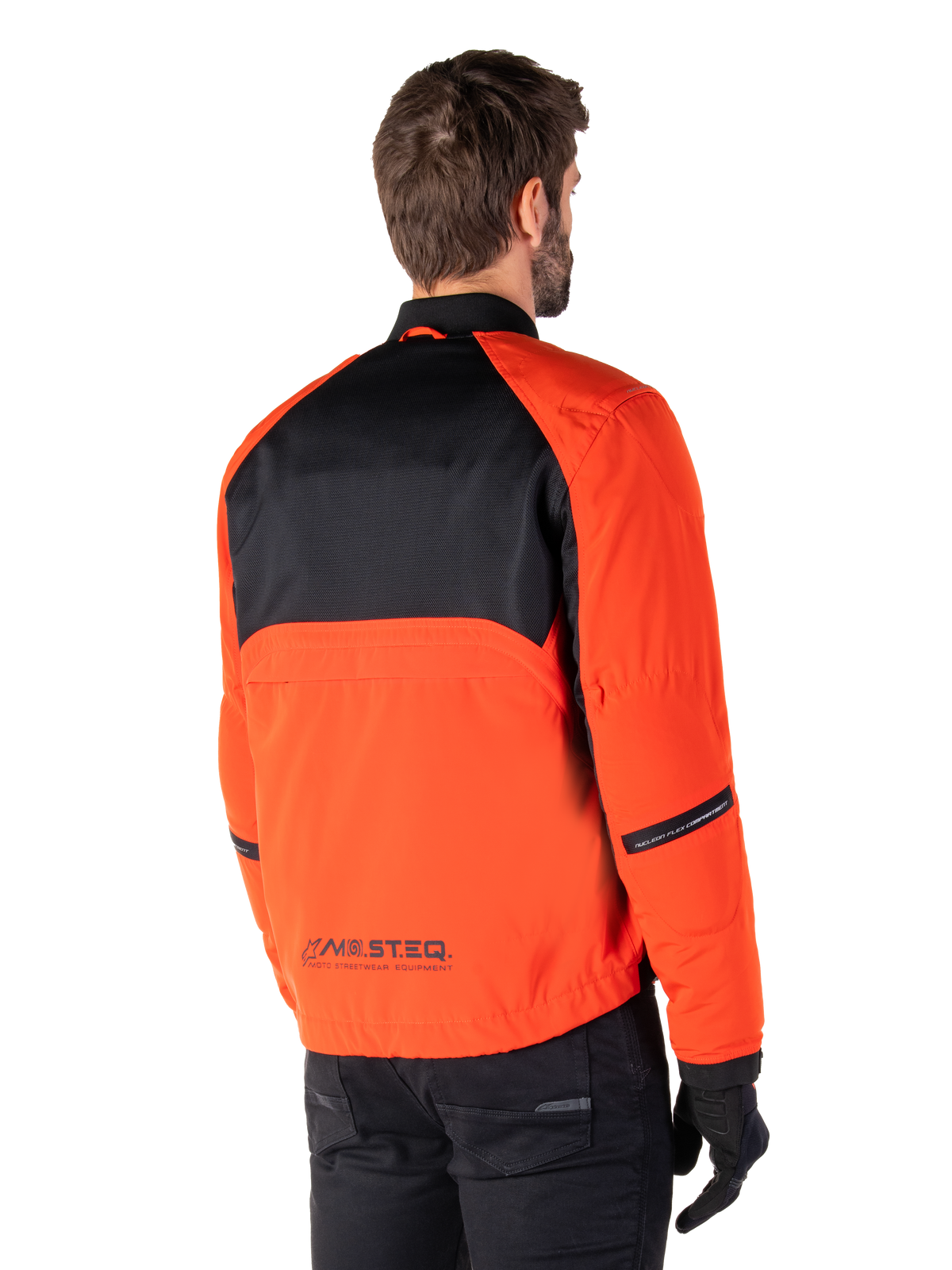 Achteraanzicht van een motorrijder die de Alpinestars Mootant Waterproof Parka draagt in Spicy Orange en zwart, voorzien van een zwart mesh paneel op de bovenrug en reflecterende MO.ST.EQ.-branding, moderne utilitaire motorjas.