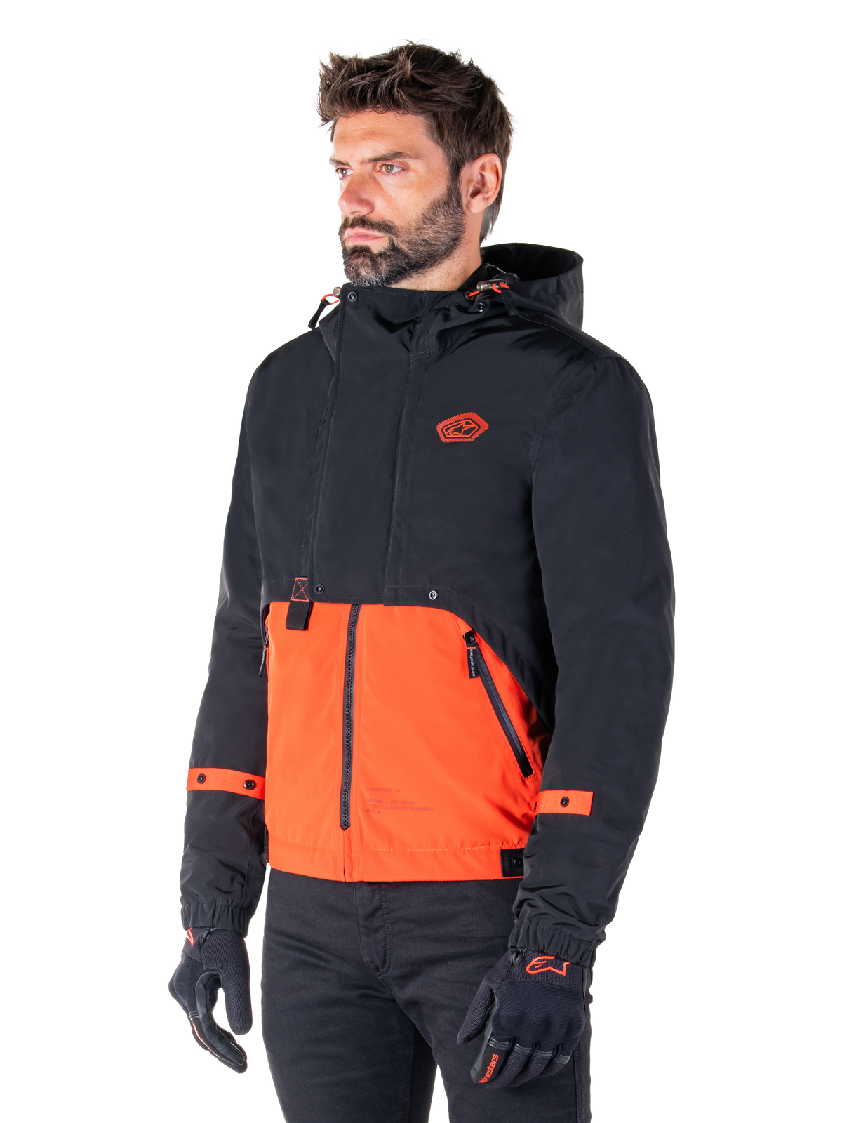 Man draagt de Alpinestars Mootant Waterproof Parka in Spicy Orange en Black, een modulaire motorjas met een zwarte bovenlaag met capuchon en een feloranje ondergedeelte, gecombineerd met zwarte motorhandschoenen en broek.