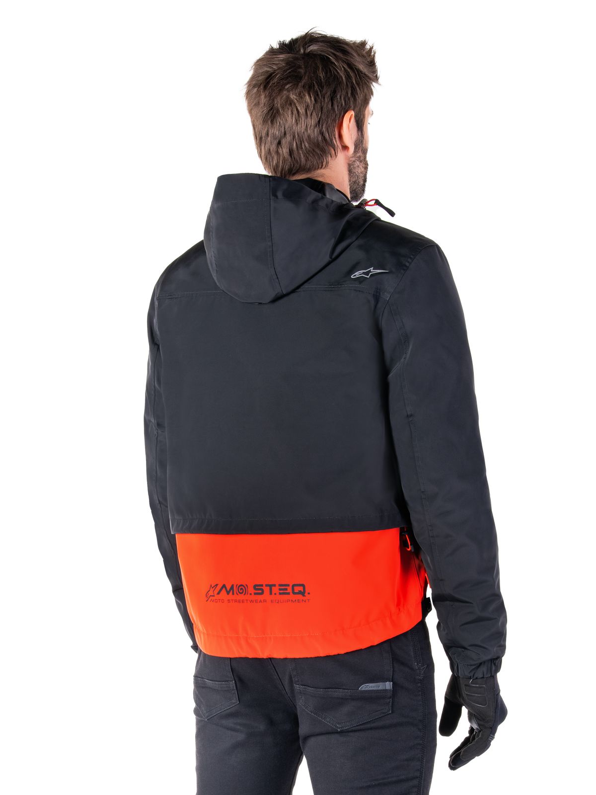 Alpinestars Mootant Waterproof Parka, Spicy Orange en zwart, achteraanzicht op een mannelijk model, voorzien van een zwart bovengedeelte met capuchon en een feloranje ondergedeelte met MO.ST.EQ.-branding, urban contemporary motorjas gedragen met een zwarte broek en handschoenen