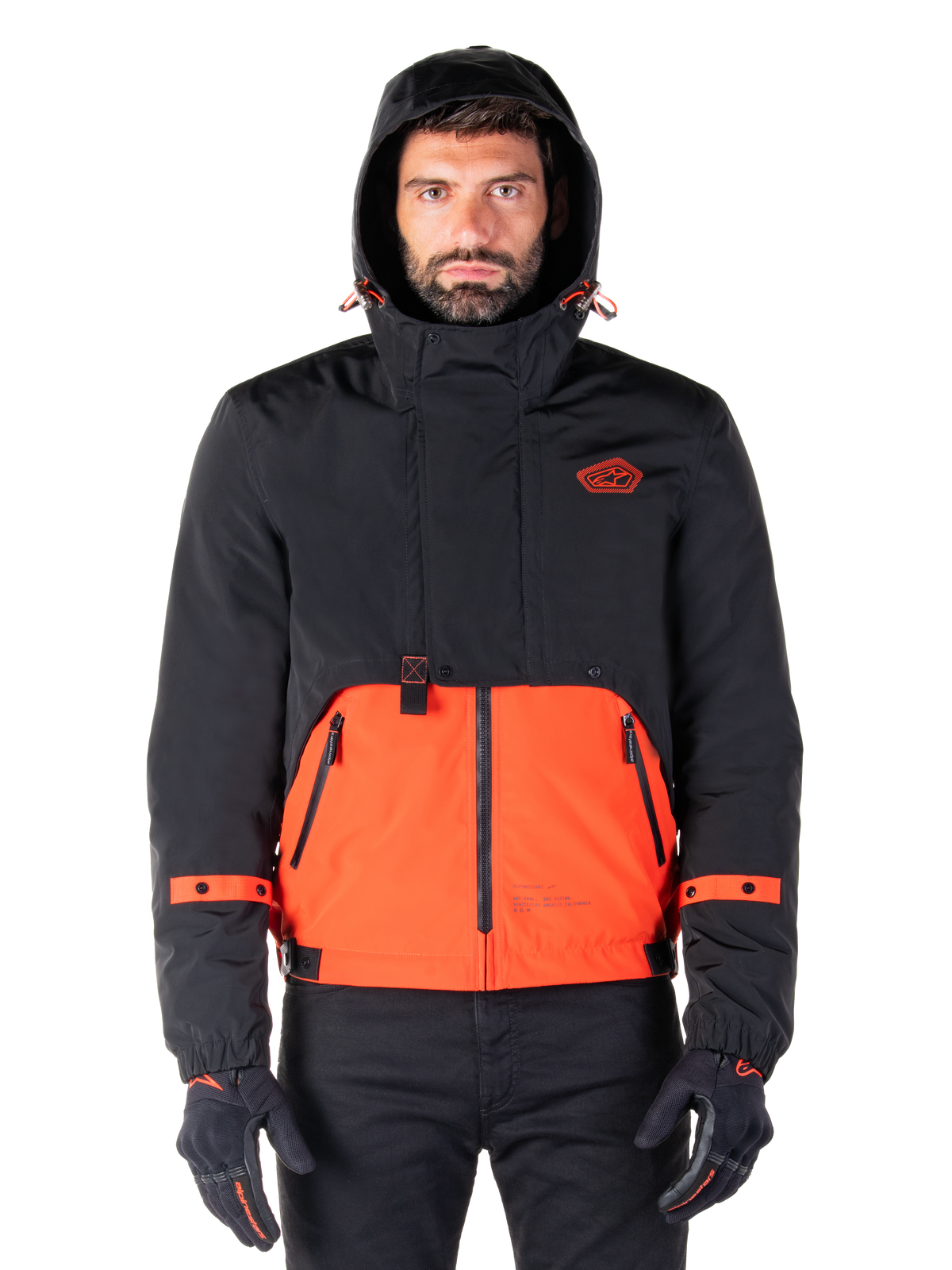 Alpinestars Mootant Waterproof Parka, motorjas, Spicy Orange en zwart, modulair all-weather ontwerp met capuchon, ritszakken aan de voorkant en oranje banddetails op de mouwen, gedragen door een model met zwarte handschoenen en broek