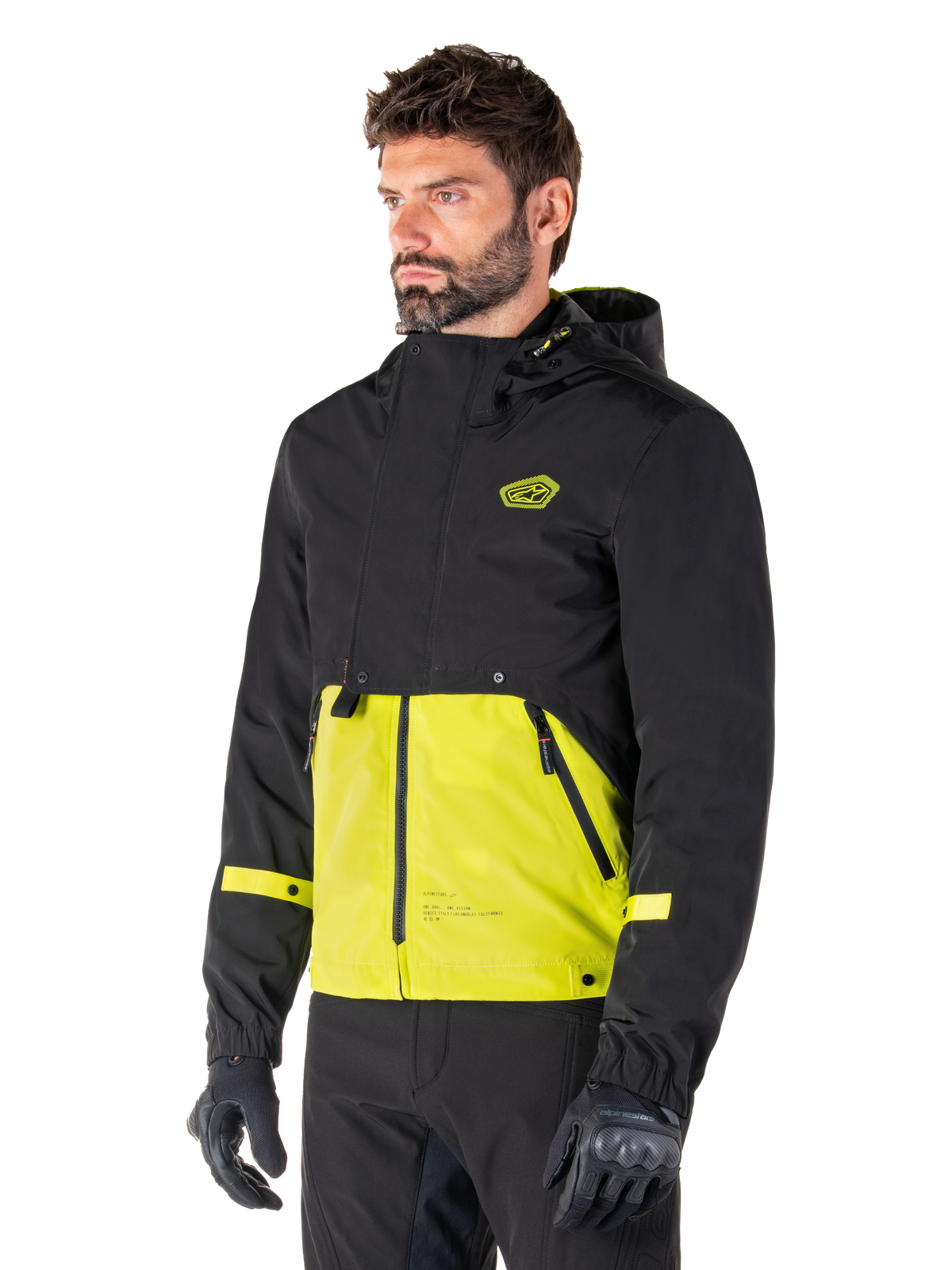Alpinestars Mootant Wasserdichter Parka in Lime Punch Black, modulare Motorradjacke getragen von einem Mann mit schwarzen Motorradhandschuhen und -hosen, schwarzer oberer Bereich und Ärmel mit leuchtend limettengrünem Unterteil, Kapuzendesign mit utilitaristischer, militärisch inspirierter Ästhetik.