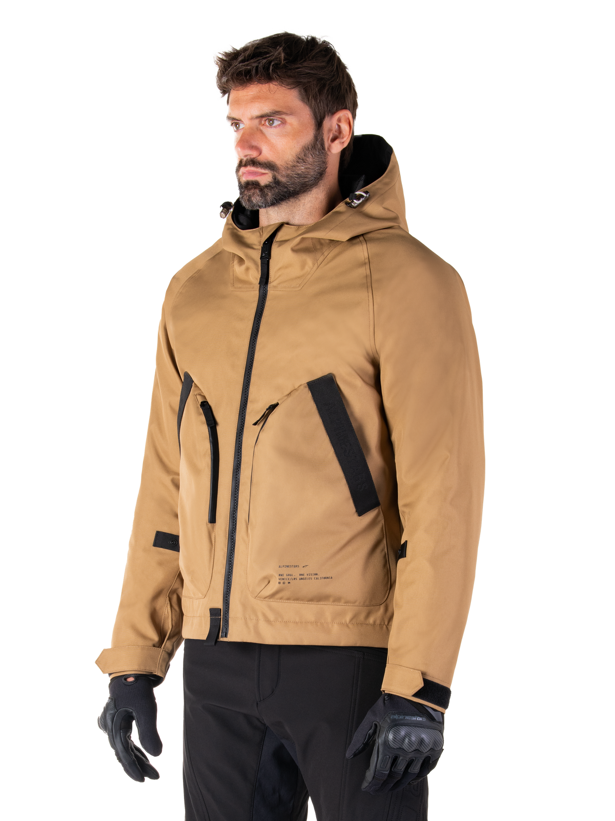 Alpinestars Morush Wasserdichter Parka, Utility Brown, männliches Model trägt Parka mit schwarzen Motorradhandschuhen und schwarzer Hose, minimalistisches, von Streetwear inspiriertes Design mit sturmsicherer Kapuze und großen Vordertaschen, wasserdichte Motorradbekleidung