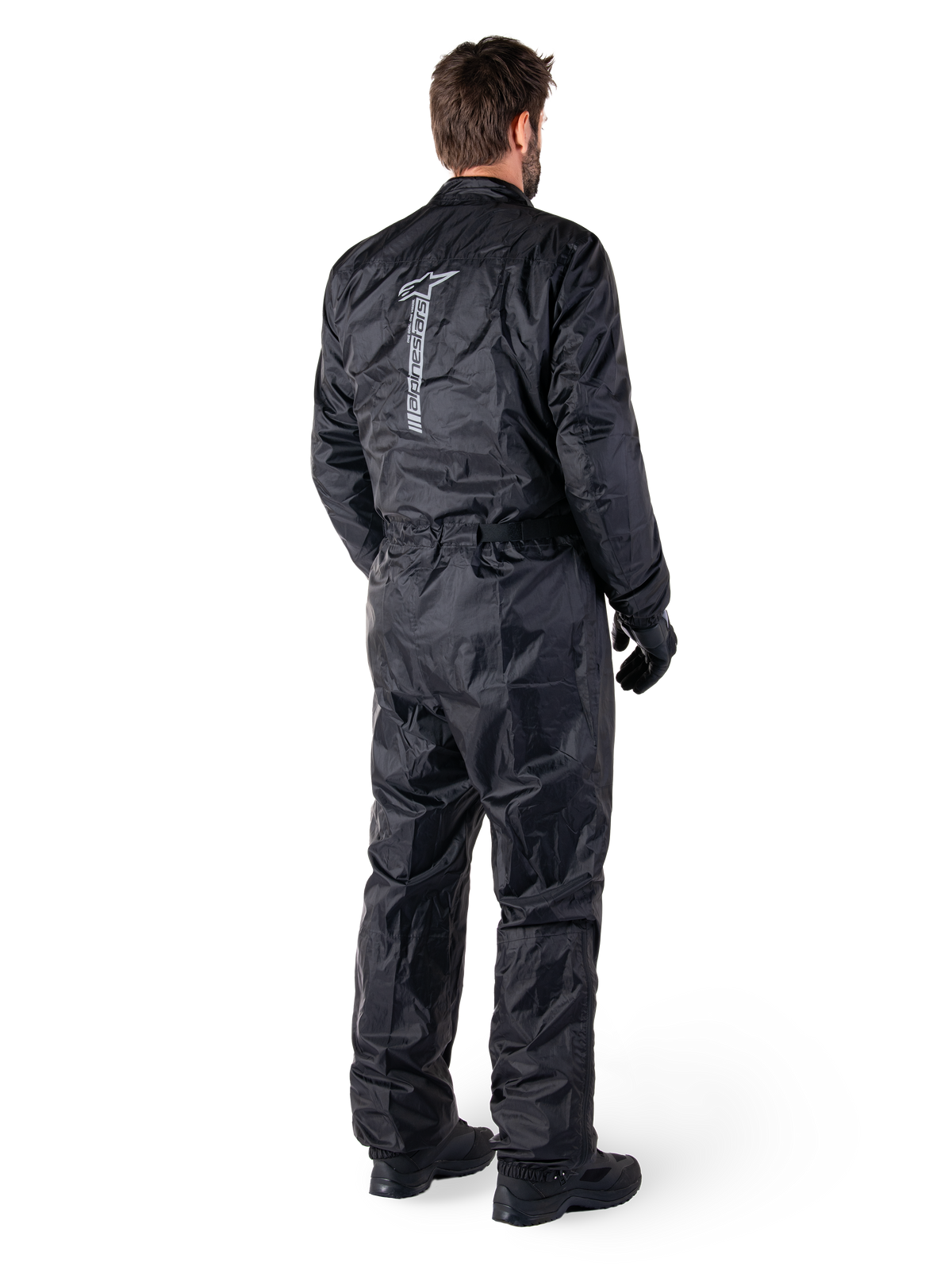 Alpinestars Hurricane Rain V2 Suit, Black one-piece waterproof motorcycle rain suit, back view with large vertical reflective logo, lightweight wind-resistant fabric, worn with black gloves and boots