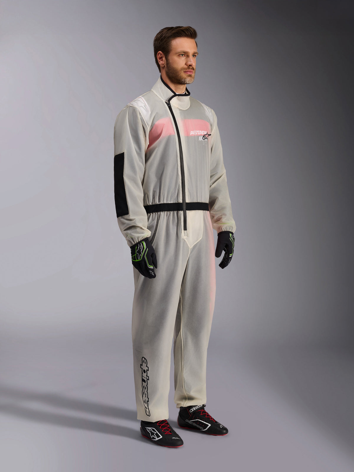 Alpinestars Kart Rain Suit, klarer wasserdichter Kart-Überanzug, halbtransparentes Material mit rosa-weißem Rennanzug darunter, schwarzer asymmetrischer Reißverschluss, verstärkter Ellbogenbesatz, schwarzer Taillenbund, schwarze Kart-Handschuhe und Rennschuhe