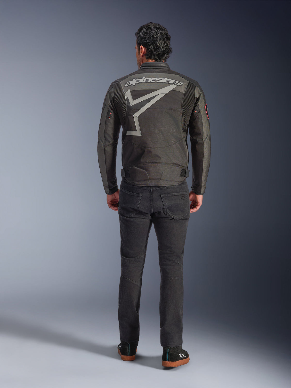 Alpinestars Axton Jacke, Motorradjacke, Schwarz Anthrazit Rot Tangerine, Rückansicht mit großem grauem Alpinestars-Logo und Sterngrafik, Akkordeon-Schulterpaneele, getragen mit schwarzer Hose und schwarzen Freizeitschuhen.