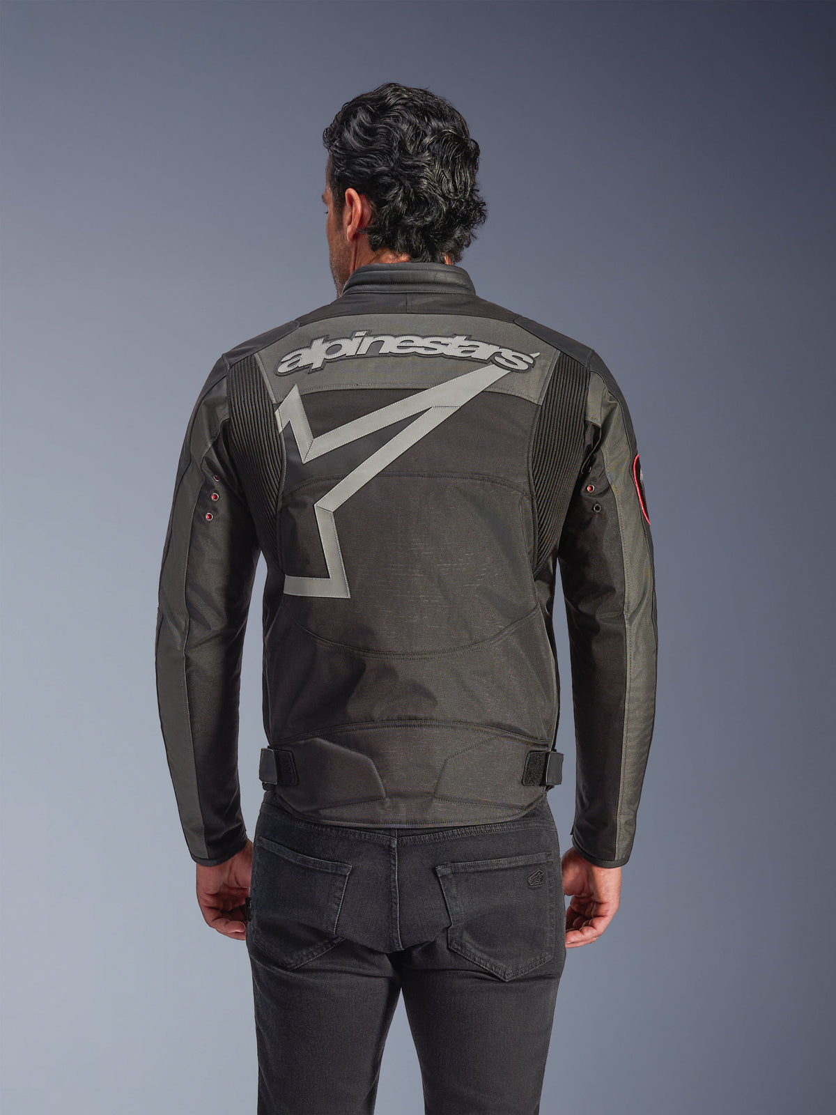 Alpinestars Axton Jacke, Motorradjacke für Herren, Schwarz Anthrazit Rot Tangerine, Rückansicht mit großem grauen Alpinestars-Schriftzug und stilisierter Sterngrafik, Akkordeon-Einsätze an den Schultern, Vintage-inspirierte Metallösen an den Ärmeln.