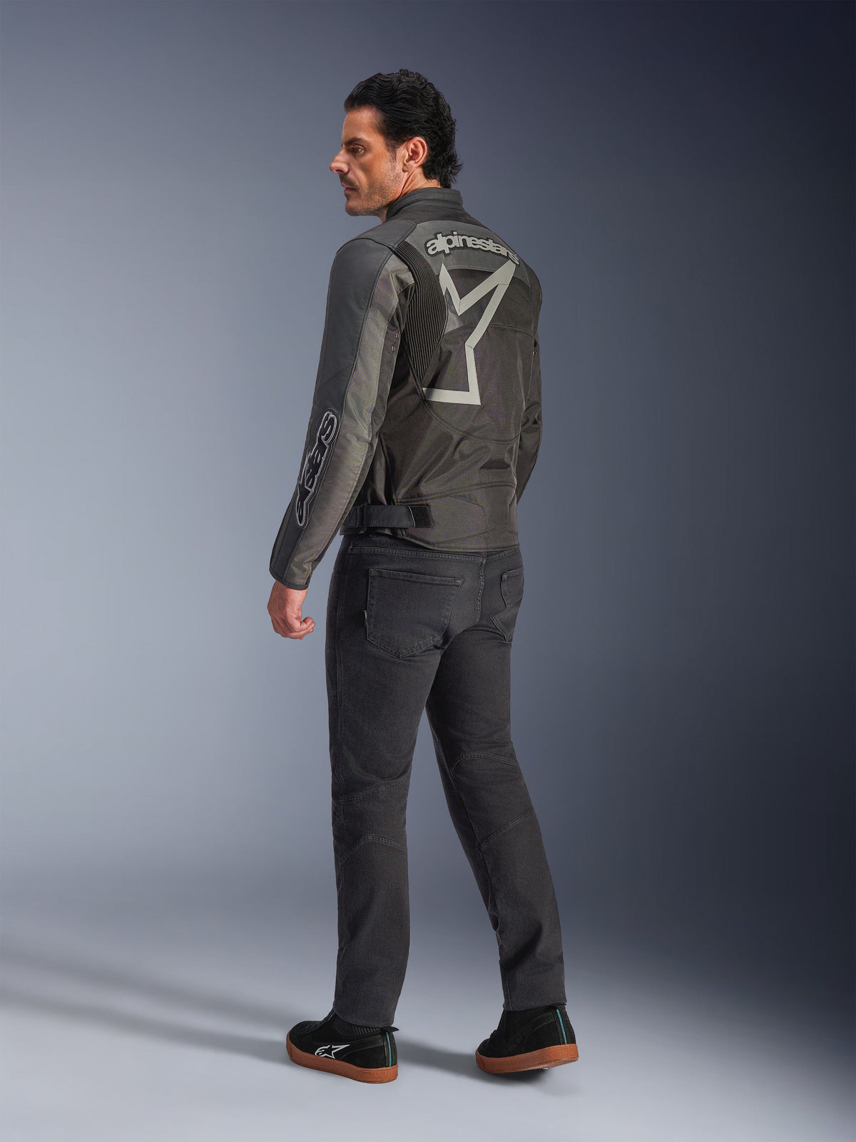 Alpinestars Axton Jacke, Motorradjacke für Herren, Schwarz und Anthrazit, Rückansicht mit einem großen grauen, umlaufenden Logo und Akkordeon-Stretcheinsätzen an den Schultern, getragen mit dunkler Fahrhose und schwarzen Sneakern, urbaner, zeitgemäßer Stil mit Racing-Tradition.
