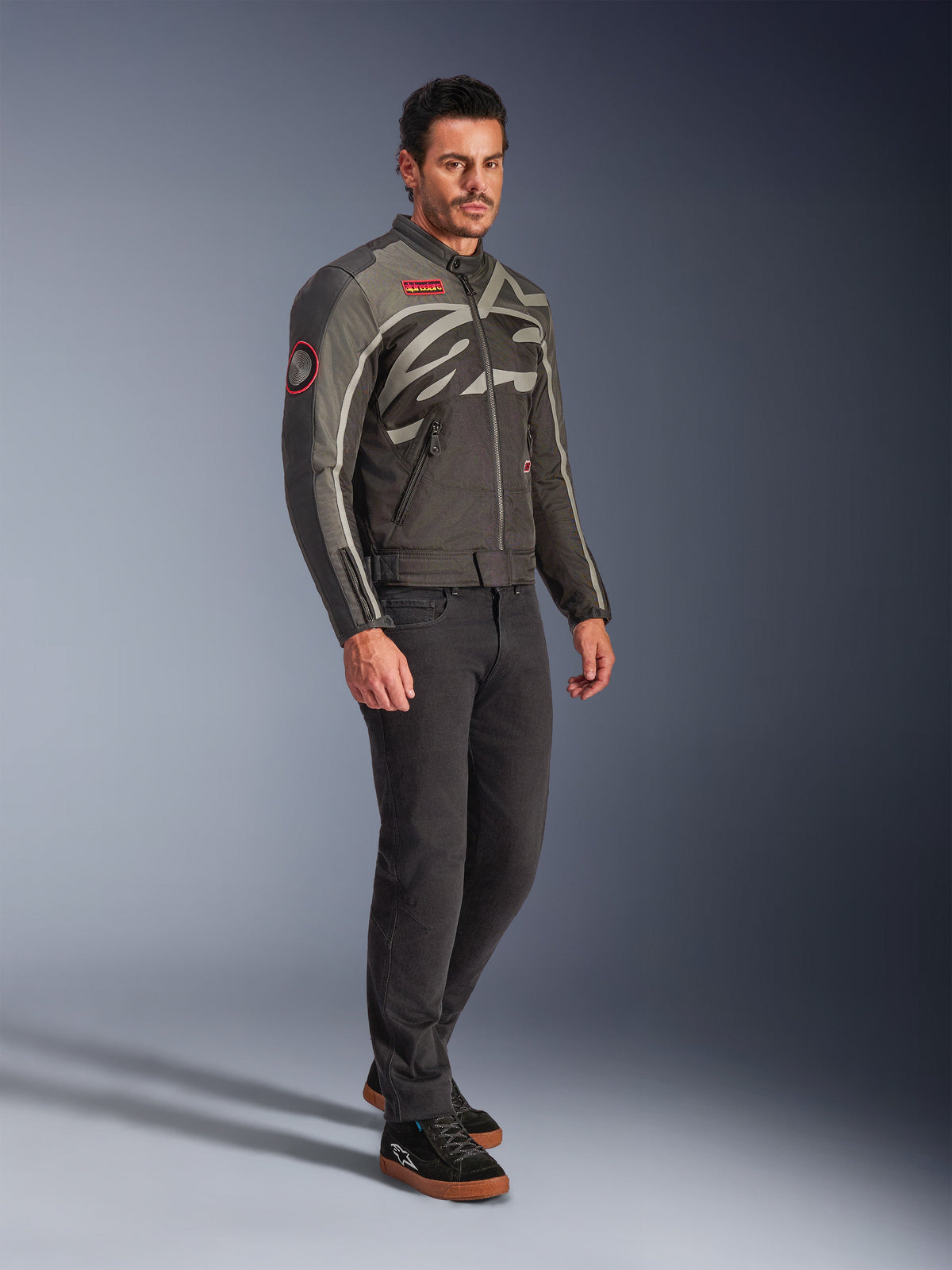 Männliches Model in der Alpinestars Axton Jacke in Black Anthracite Red Tangerine, Vintage-inspirierte Motorradjacke mit umlaufendem Logo und roten Akzenten, kombiniert mit schwarzer Hose und schwarzen Motorrad-Sneakern, urbane zeitgemäße Motorradausrüstung