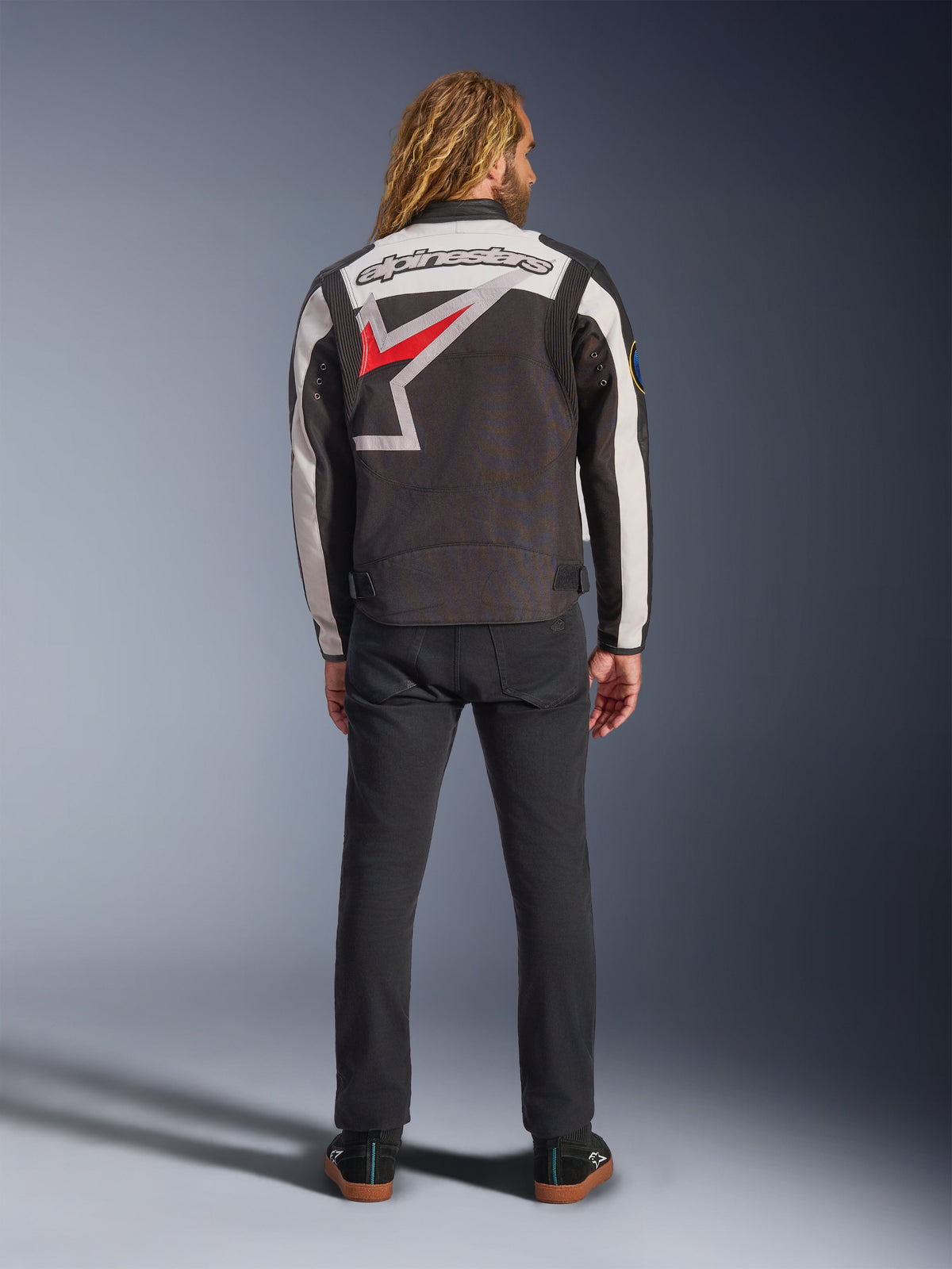 Veste Alpinestars Axton, veste de moto, noir, blanc cassé et rouge vif, vue de dos sur un mannequin, avec un grand logo étoile Alpinestars dans le dos, empiècements de manches blanc cassé, portée avec un pantalon noir, design inspiré des vêtements d'équipe de course vintage