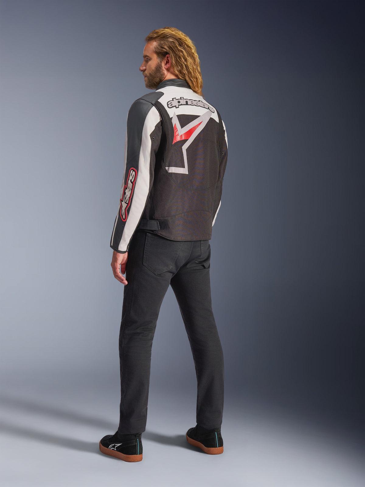 Veste Alpinestars Axton, veste de moto, noir, blanc cassé et rouge vif, vue arrière portée par un mannequin homme, arborant un grand logo étoile et le nom de la marque Alpinestars dans le dos, design d'inspiration vintage racing avec des panneaux en accordéon, associée à un pantalon noir et des baskets