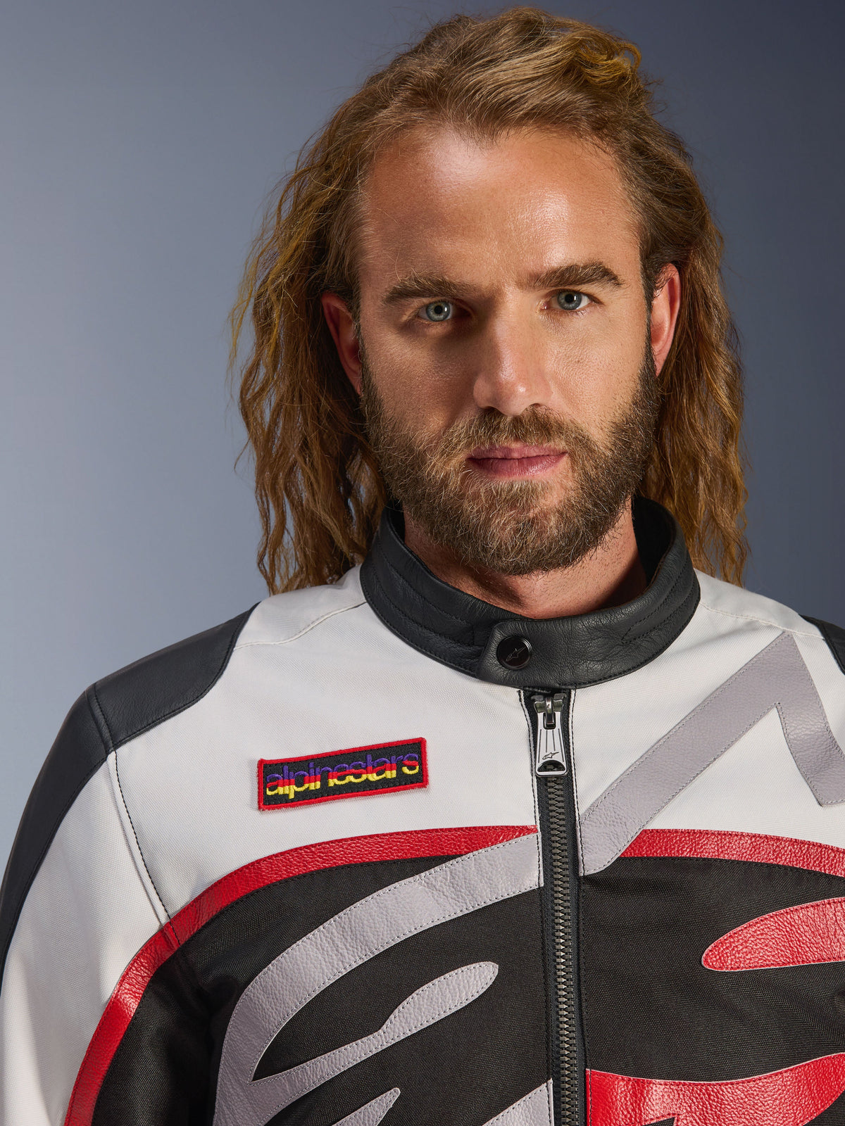 Homme portant la veste Alpinestars Axton, veste de moto d'inspiration vintage en Noir, Blanc Cassé et Rouge Vif, vue rapprochée montrant l'écusson de poitrine de la marque et le col de motard minimaliste, équipement de route contemporain