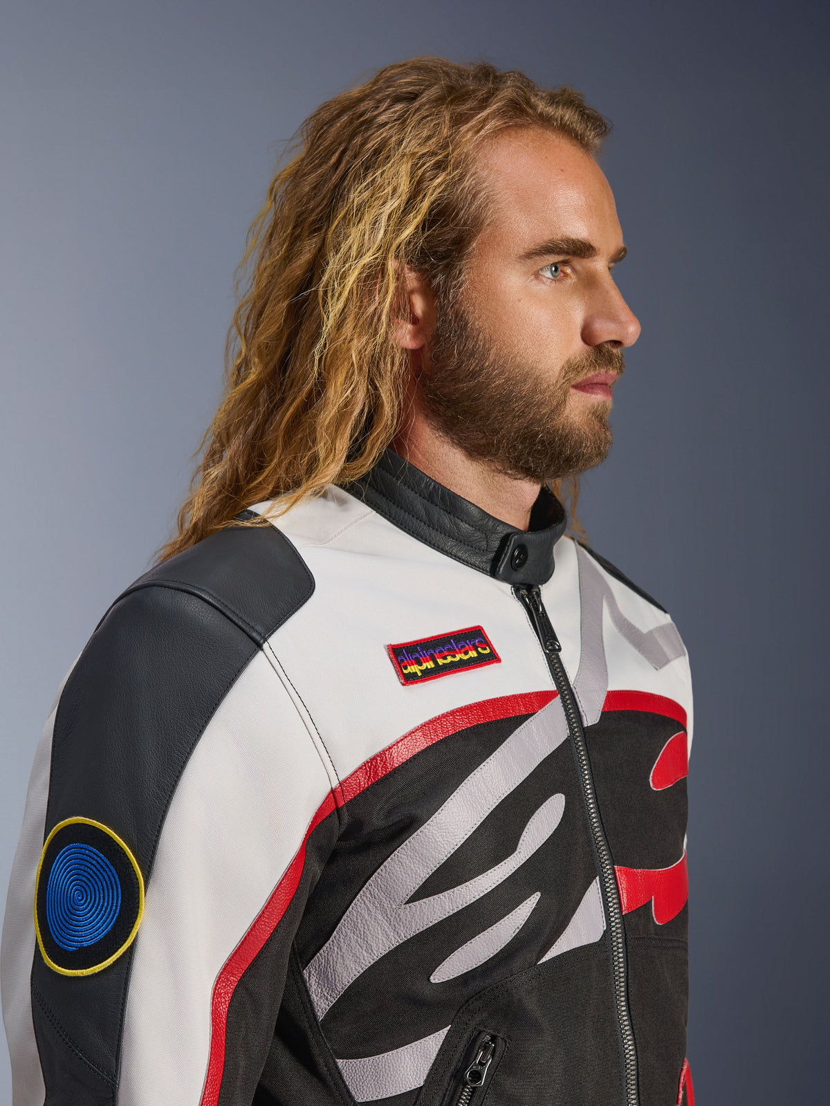 Homme aux cheveux longs et barbu portant la veste Alpinestars Axton, veste de moto, noir, blanc cassé et rouge vif, design d'inspiration racing vintage avec écussons de la marque et logo enveloppant, vue de profil rapprochée, style urbain contemporain