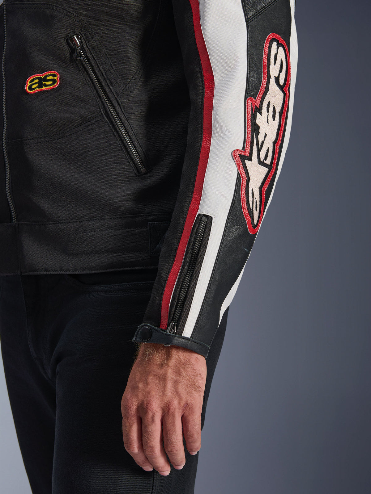 Veste Alpinestars Axton, gros plan de la manche et du buste, noir avec des accents blanc cassé et rouge vif, avec un grand logo Alpinestars brodé sur le bras, des bandes de course verticales, un poignet zippé avec bouton-pression et une construction en textile robuste.