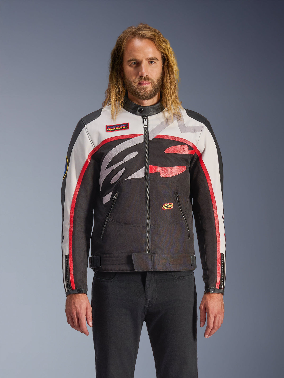 Alpinestars Axton Jacket, veste de moto, noir, blanc cassé et rouge vif, avec des motifs abstraits audacieux sur la poitrine et un logo enveloppant, manches à rayures rouges, design d'inspiration vintage avec un col minimaliste, portée par un mannequin avec un jean noir.