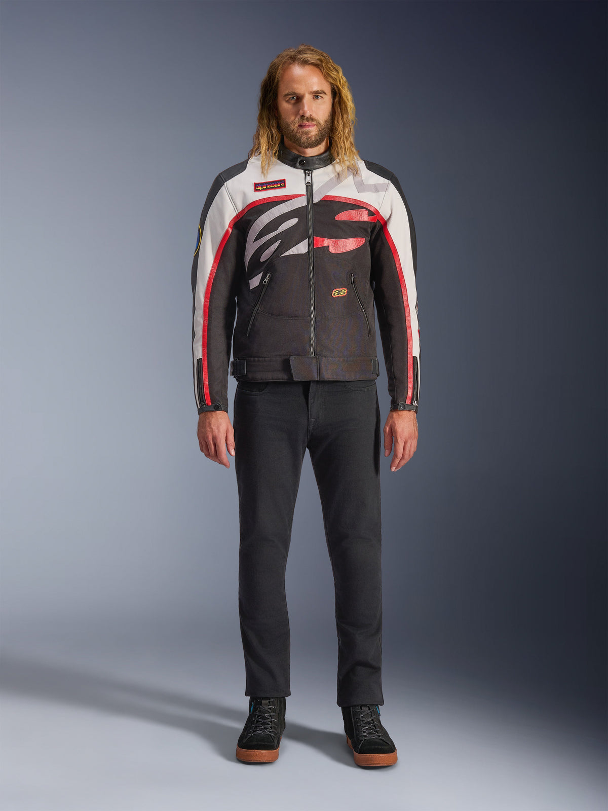 Veste Alpinestars Axton, veste de moto, noir, blanc cassé et rouge vif, portée par un mannequin avec un pantalon noir et des baskets, dotée d'un logo enveloppant audacieux et d'un design inspiré de la course vintage