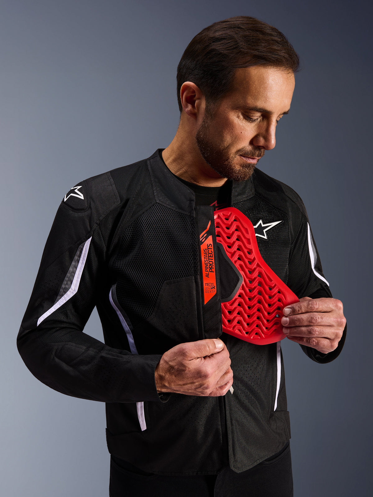 Alpinestars AST V3 Air Jacket in het zwart, man toont een felrode borstprotector met honingraatpatroon, benadrukt Tech-Air Ready compatibiliteit en geïntegreerde interne protectie voor motorveiligheid.