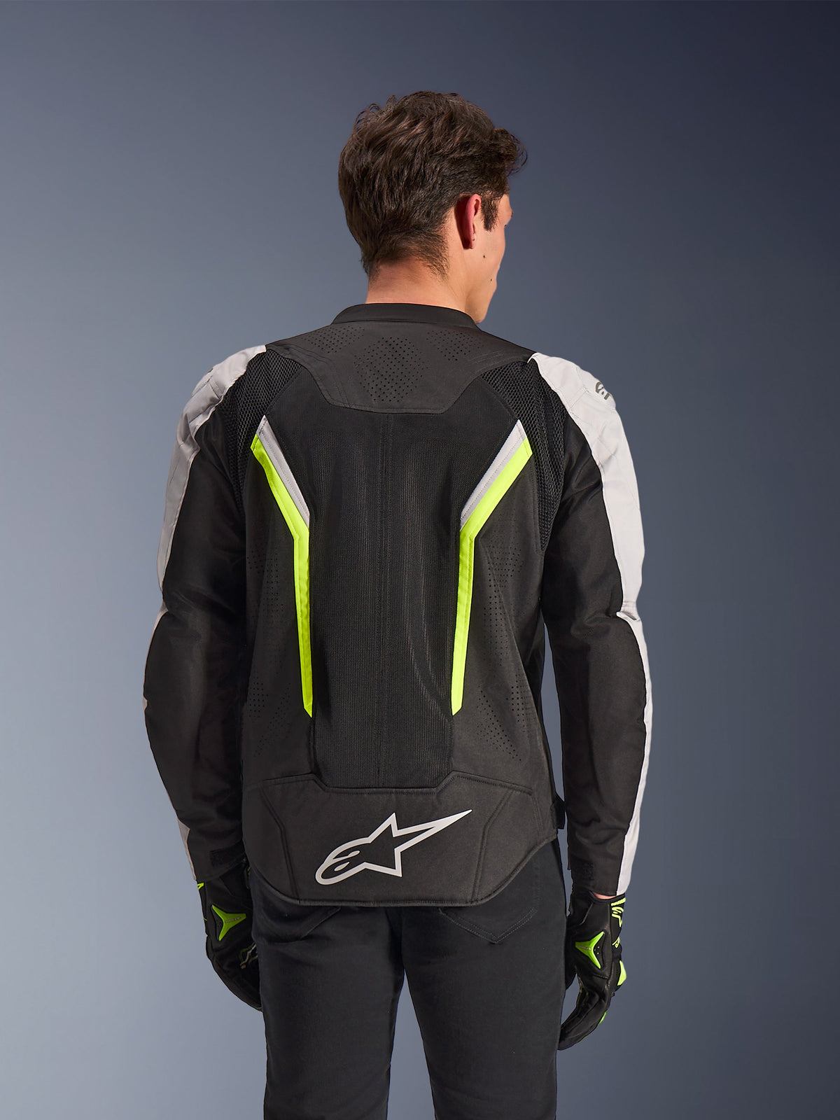 Veste Alpinestars AST V3 Air, Noir Argent Jaune Fluo, vue arrière sur modèle, veste de moto en textile dotée de panneaux de ventilation en maille double technologie, bandes verticales jaune fluo, manches argentées et logo Alpinestars blanc sur le bas du dos.