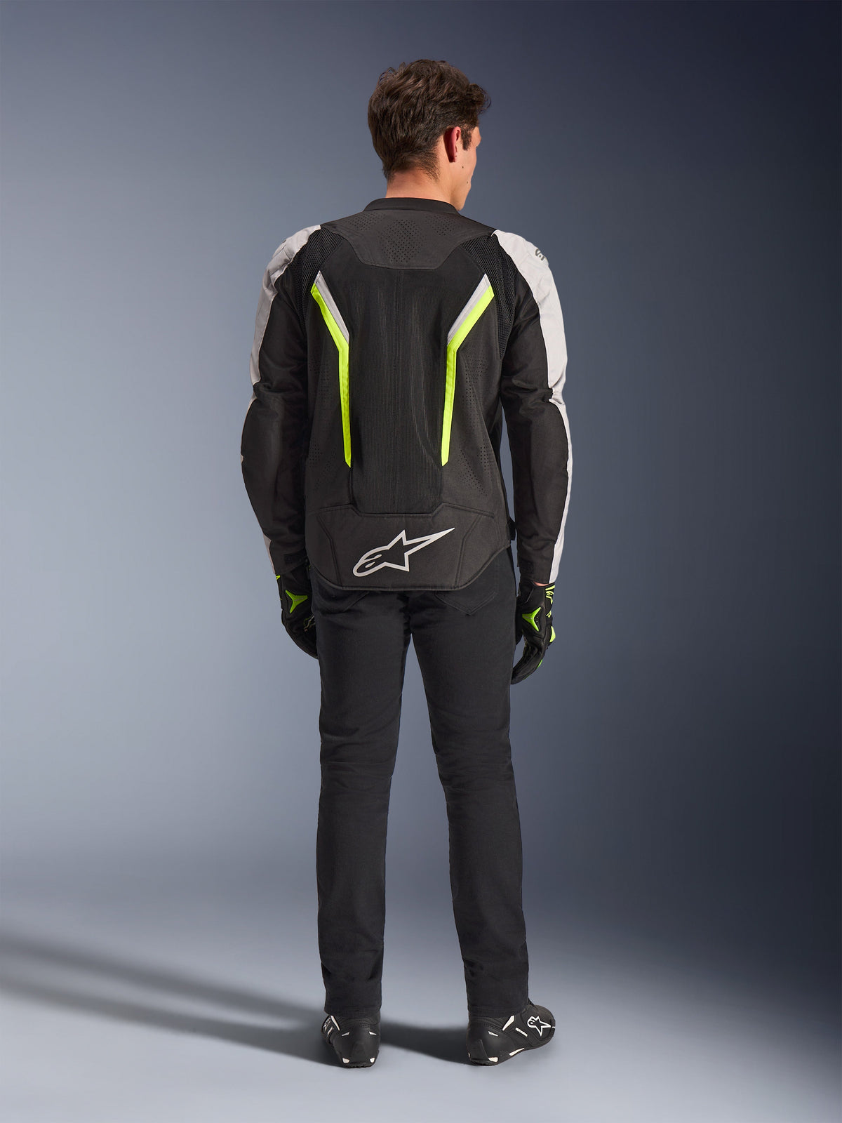 Veste Alpinestars AST V3 Air, veste de moto en textile, Noir, Argent et Jaune Fluo, vue arrière montrant les panneaux de ventilation en maille et les accents fluorescents, portée avec un pantalon noir et des gants de moto, équipement de conduite sportive haute performance