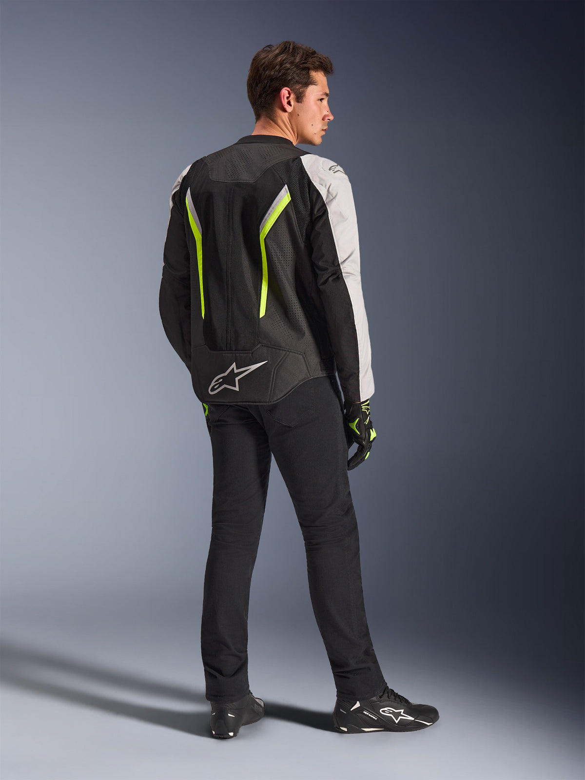 Veste Alpinestars AST V3 Air en Noir Argent Jaune Fluo, veste de moto en textile portée par un pilote vu de dos, présentant de larges panneaux en mesh et des accents jaune fluo, assortie à un pantalon, des gants et des chaussures de moto noirs, équipement technique pour la conduite sportive.