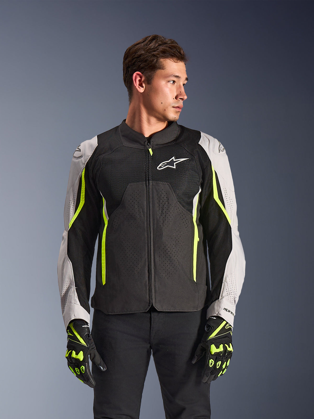 Alpinestars AST V3 Air Jacket, veste de moto en textile, noire et argentée avec des accents jaune fluo, dotée de larges panneaux de ventilation en mesh et de perforations au laser, portée par un modèle avec des gants assortis, équipement de performance sportive.