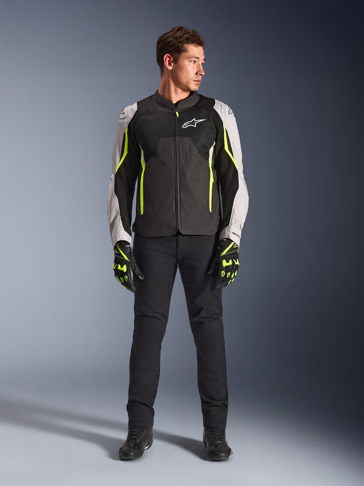 Modèle masculin portant la veste Alpinestars AST V3 Air en Noir Argent Jaune Fluo, dotée de panneaux de ventilation en mesh et d'accents fluorescents, associée à un pantalon de moto noir et des gants assortis, équipement de performance pour conduite sportive.