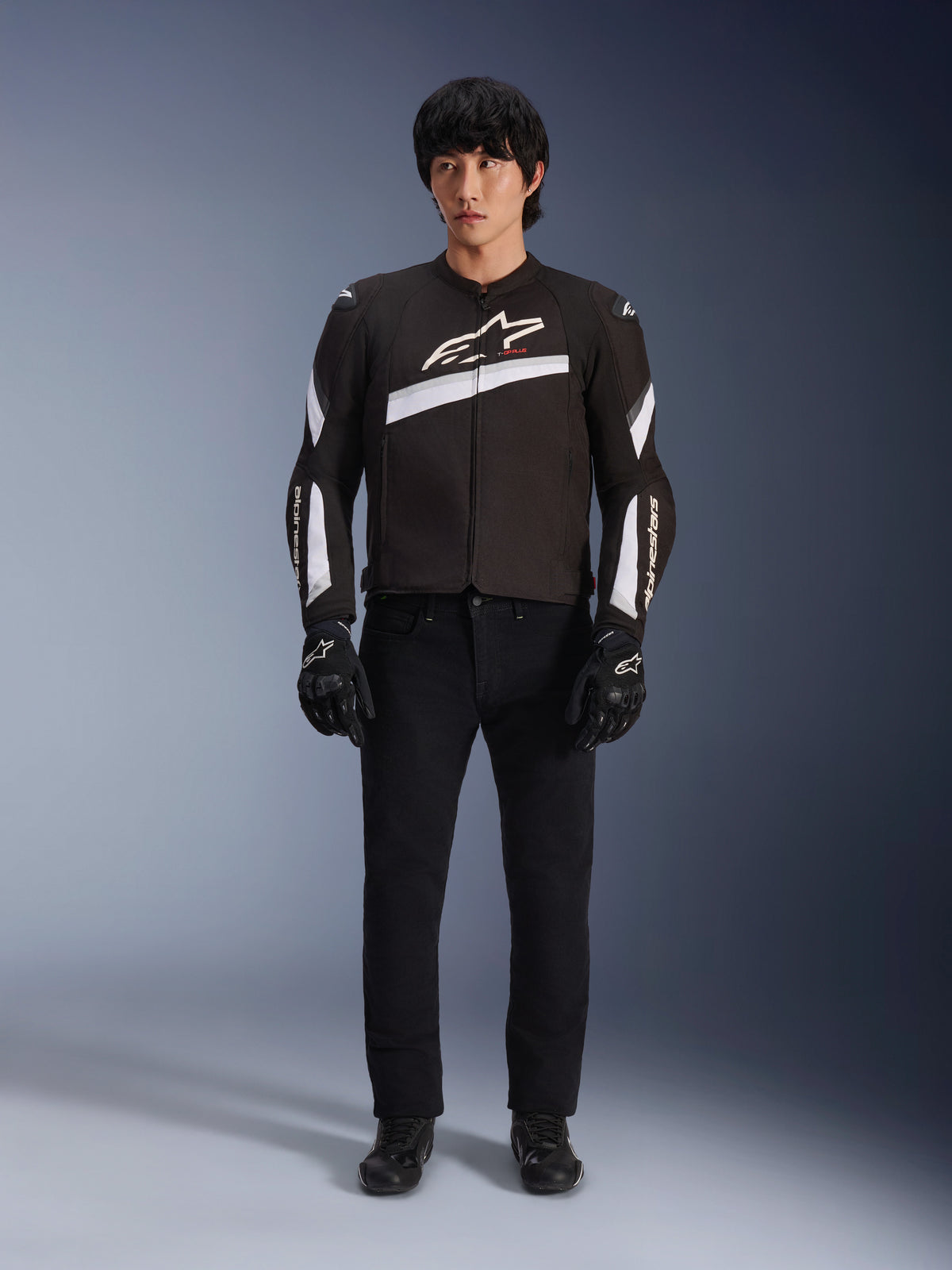 Alpinestars T-GP Plus R V4 Jacket, zwart en wit, sportieve textiele motorjas, gedragen door een model met zwarte broek en handschoenen, voorzien van een opvallend wit Alpinestars-logo op de borst en mouwen, strak op racen geïnspireerd ontwerp