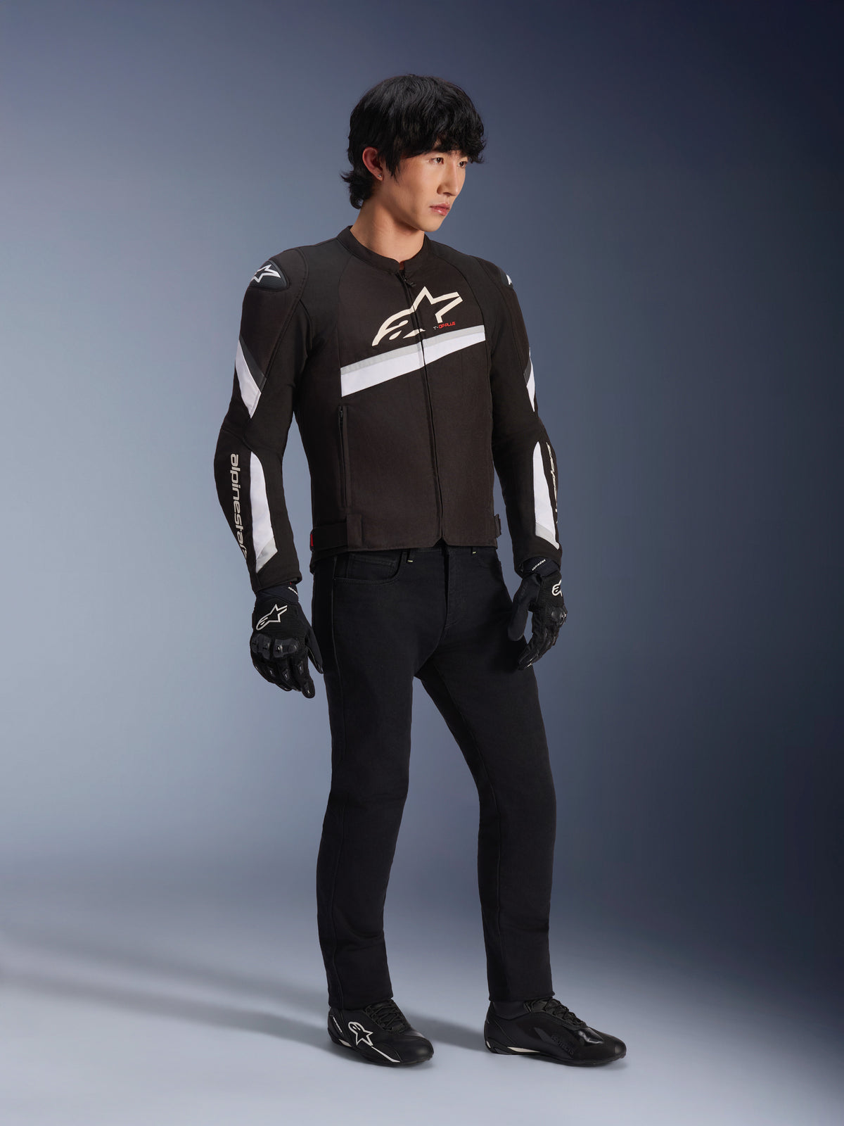 Alpinestars T-GP Plus R V4 Jas, zwart-wit textiele motorjas gedragen door een model met zwarte motorbroek en handschoenen, voorzien van witte Alpinestars-logo's op de borst en mouwen, sportief ontwerp voor wegprestaties