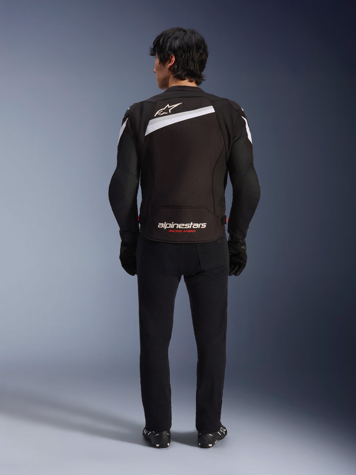 Alpinestars T-GP Plus R V4 Jacket, zwart-witte sportieve textiele motorjas, achteraanzicht met wit Alpinestars-logo en 'RACING AHEAD' branding, gedragen met zwarte broek en laarzen, gestroomlijnd ontwerp voor sportieve rijprestaties.