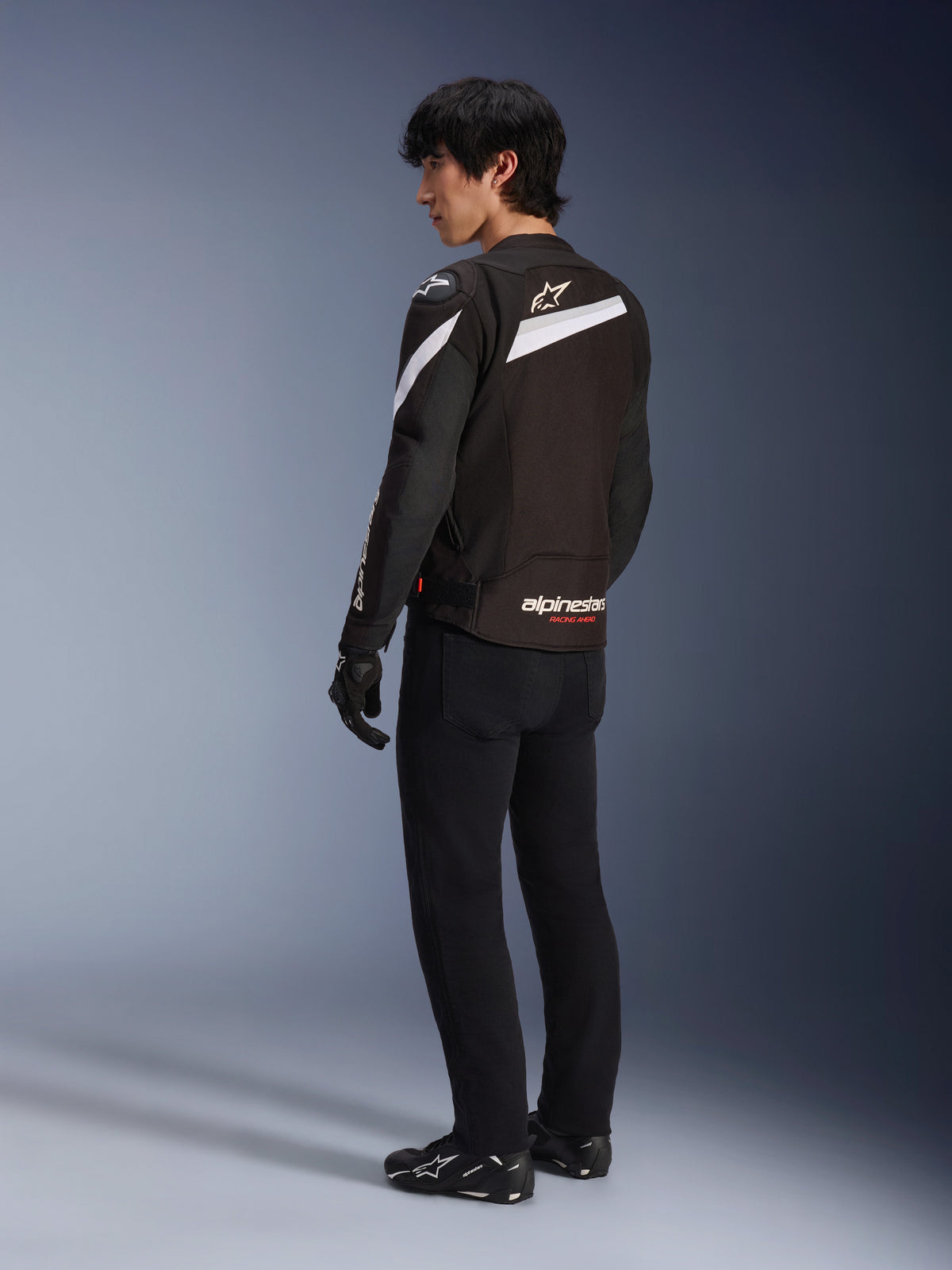 Mannelijke motorrijder die de Alpinestars T-GP Plus R V4 Jas in zwart en wit draagt, achteraanzicht met witte diagonale accenten en branding op de onderrug, gecombineerd met een zwarte motorbroek, handschoenen en technische laarzen, sportieve textiele motoruitrusting
