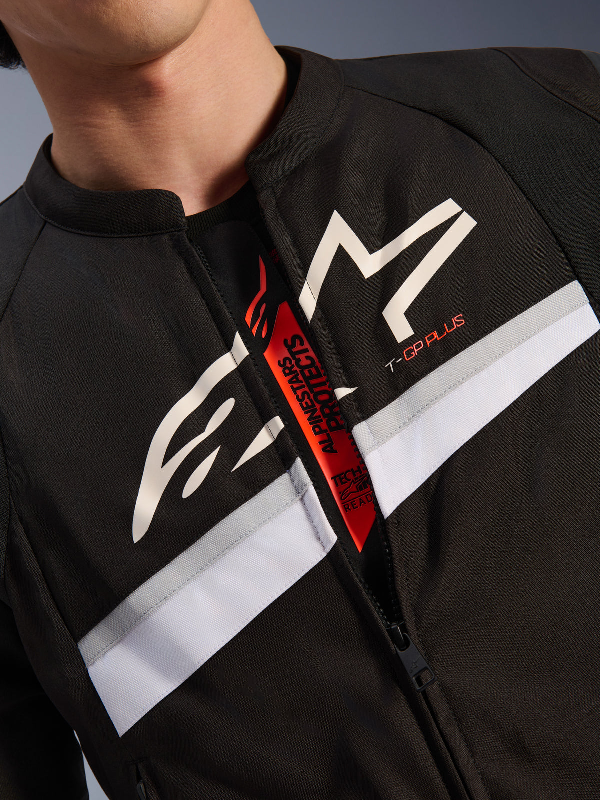 Alpinestars T-GP Plus R V4 jas, close-up van borst en nek, zwart textiel met wit sterlogo en T-GP PLUS-branding, witte en grijze horizontale streep, open rits onthult rode binnentab met Tech-Air Ready-tekst