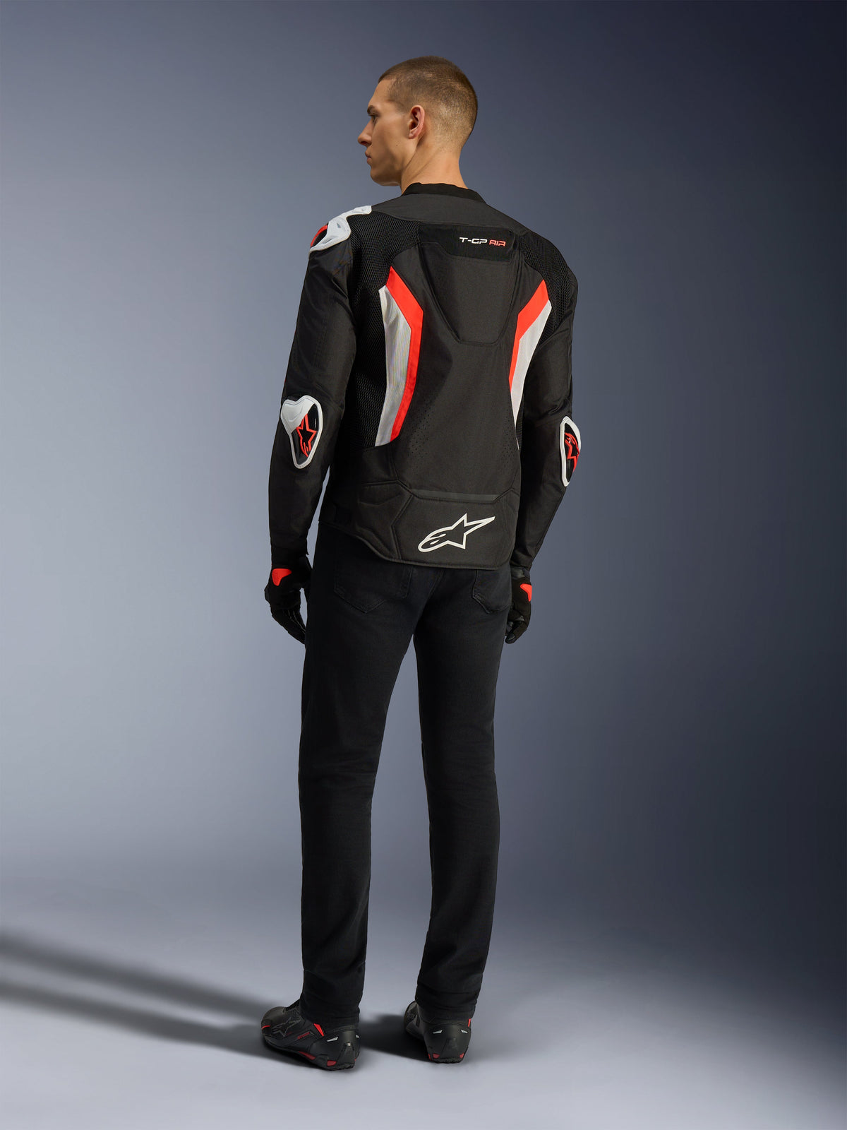 Veste Alpinestars T-GP Air, blouson de moto noir rouge fluo, vue arrière sur un modèle, avec panneaux de ventilation en maille, bosse aérodynamique et protections externes blanches aux coudes et aux épaules, porté avec un pantalon noir et des chaussures de moto