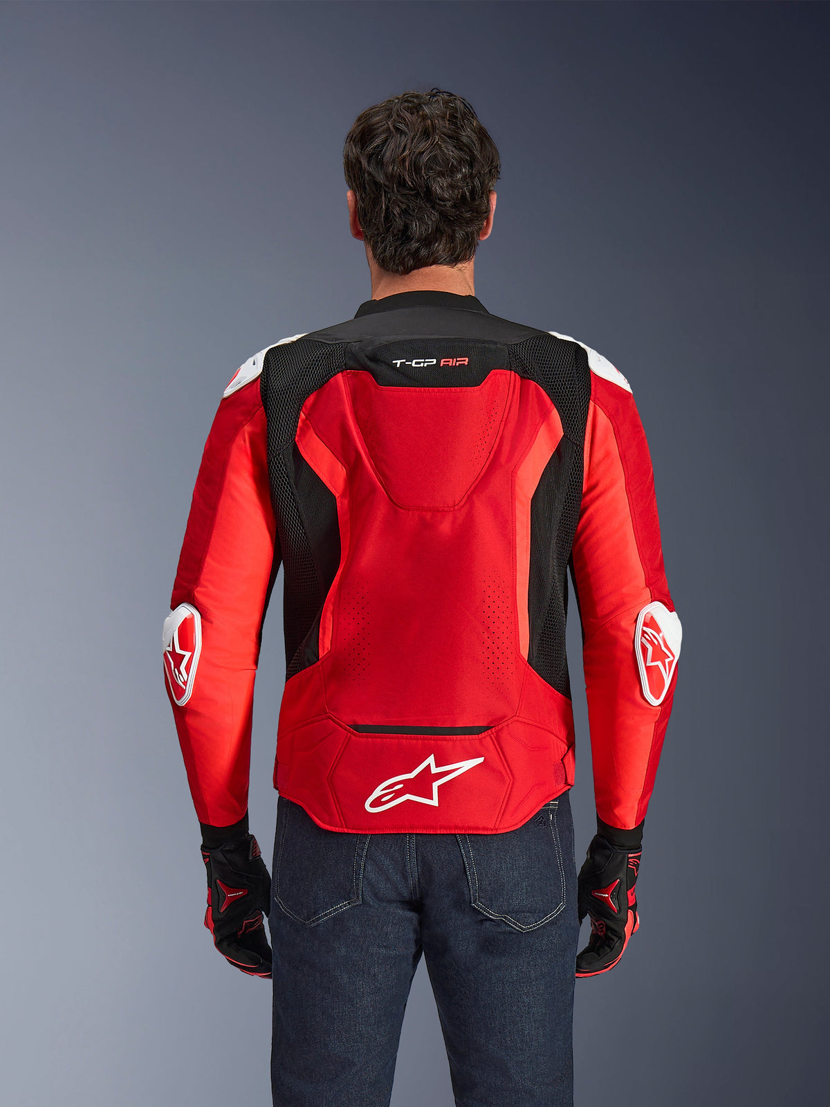 Alpinestars T-GP Air Jacke, Textil-Motorradjacke, Leuchtendes Rot und Schwarz mit weißen Akzenten, Rückansicht mit Aero-Höcker und Mesh-Belüftungseinsätzen, Alpinestars Stern-Logo und T-GP Air Branding, getragen mit dunkler Jeans und Rennhandschuhen.