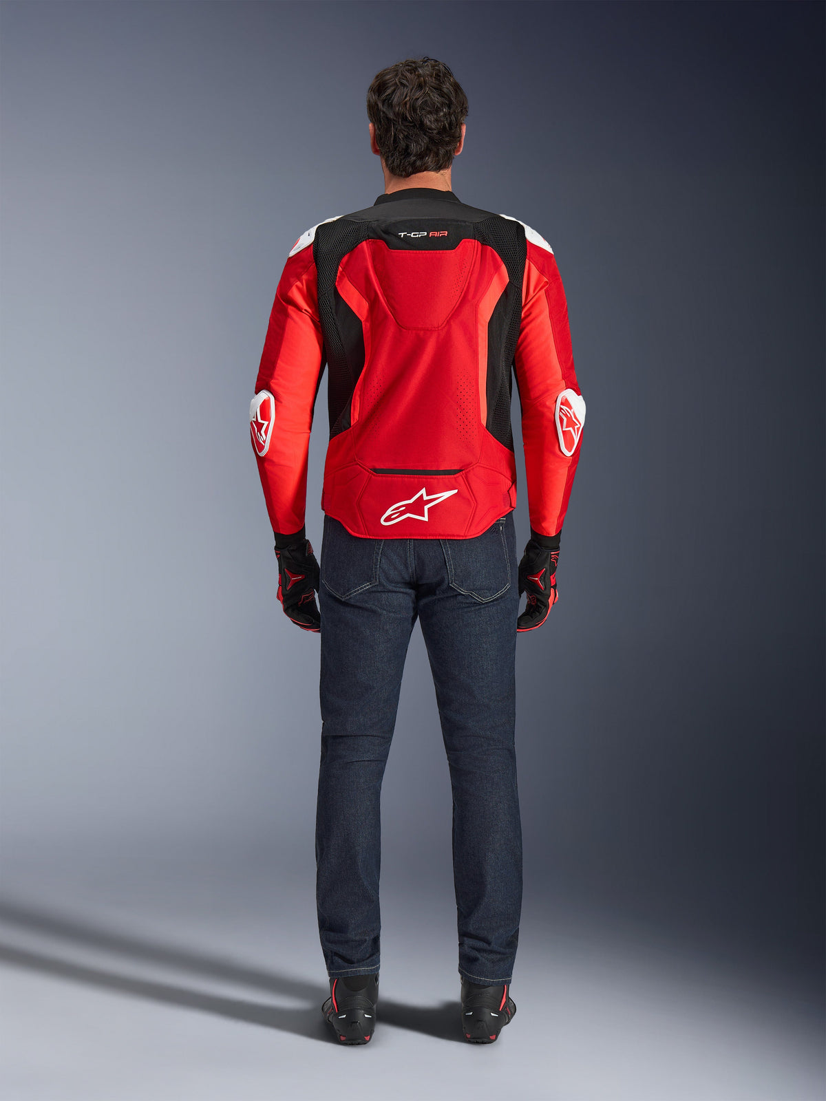 Alpinestars T-GP Air Jacke, Rückansicht, Hellrot und Schwarz, mit weißem Logo am unteren Rücken und 'T-GP AIR'-Branding, getragen mit dunkelblauen Denim-Jeans und schwarz-roten Motorradhandschuhen, sportliche Performance-Motorradausrüstung
