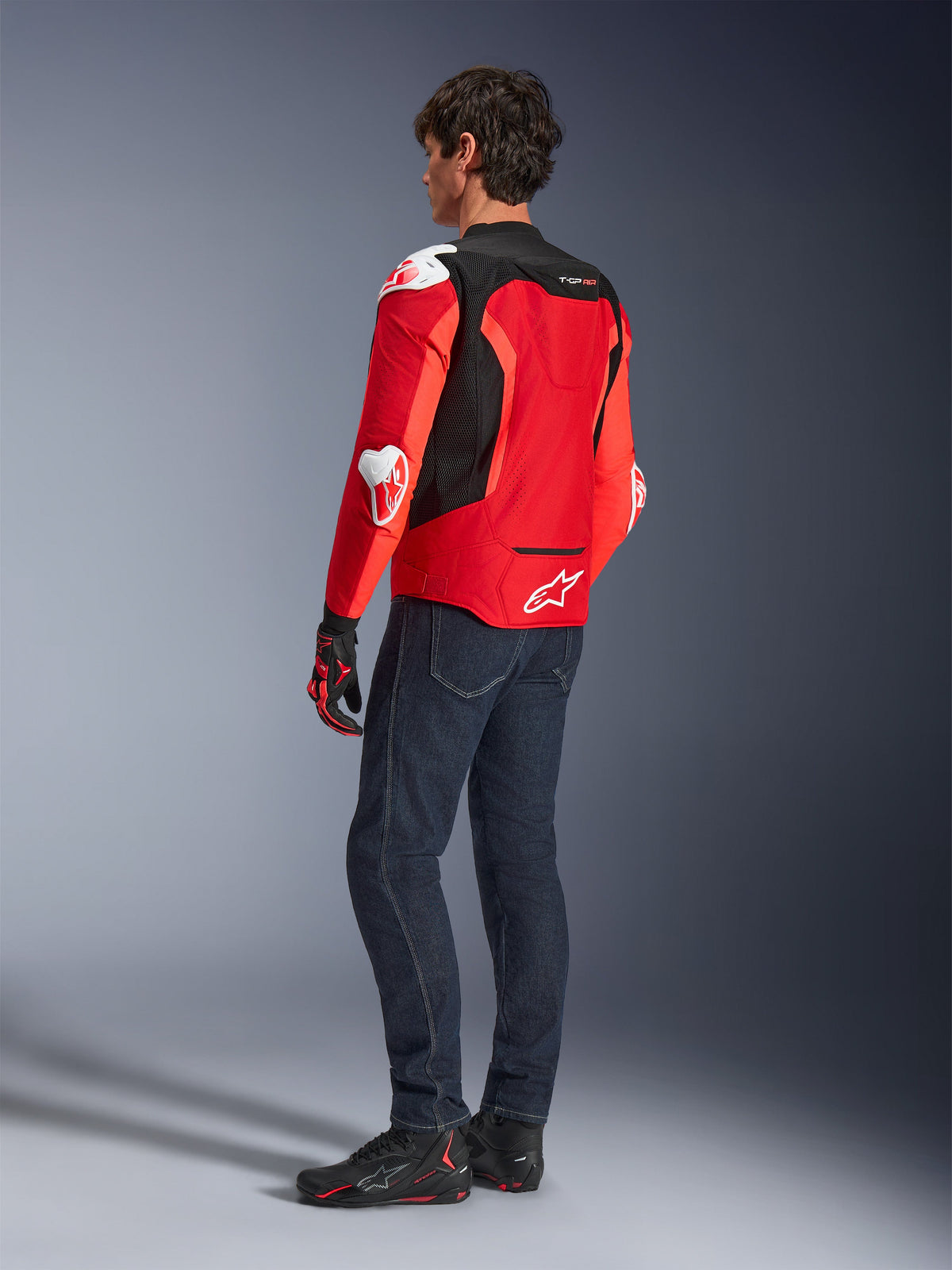 Alpinestars T-GP Air Jacke, Rückansicht eines Mannes in einer leuchtend roten und neongroten Textiljacke mit schwarzen Mesh-Einsätzen und weißen Protektoren-Schleifern, kombiniert mit dunkelblauen Jeans und schwarzen Motorradschuhen, Sport-Motorradausrüstung.