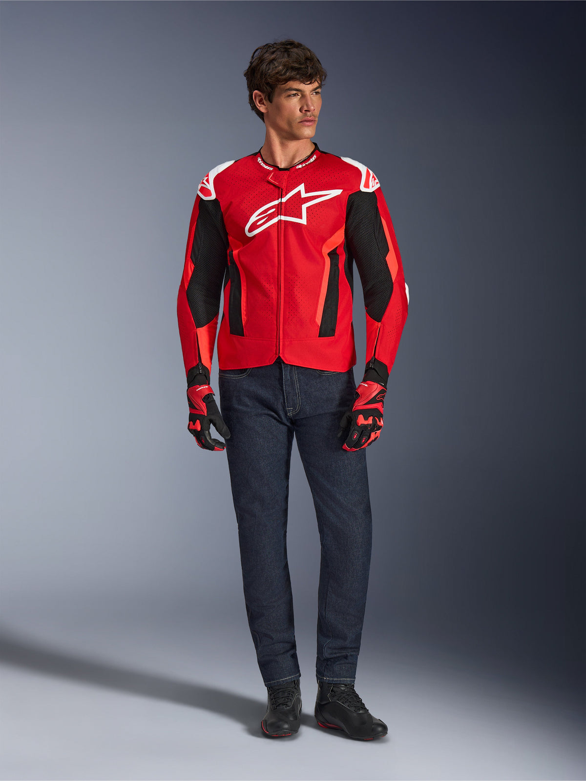 Alpinestars T-GP Air Jacke, Hellrot Fluo-Rot Schwarz, männliches Model in kompletter Motorradausrüstung mit dunkelblauer Jeans und passenden Handschuhen, Sport-Textiljacke mit perforierten Mesh-Einsätzen und weißem Astars-Logo, Studioaufnahme.