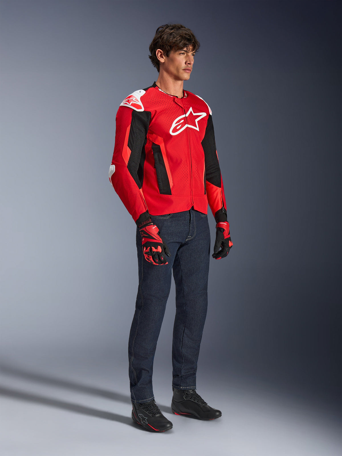 Alpinestars T-GP Air Jacke, sportliche Motorradjacke, leuchtendes Rot und Schwarz mit weißen Akzenten, getragen von einem Model mit dunkelblauen Jeans und roten Rennhandschuhen, mit High-Flow-Mesh-Einsätzen und weißen Schulterschleifern, aerodynamische Sport-Passform für Fahrten bei warmem Wetter.