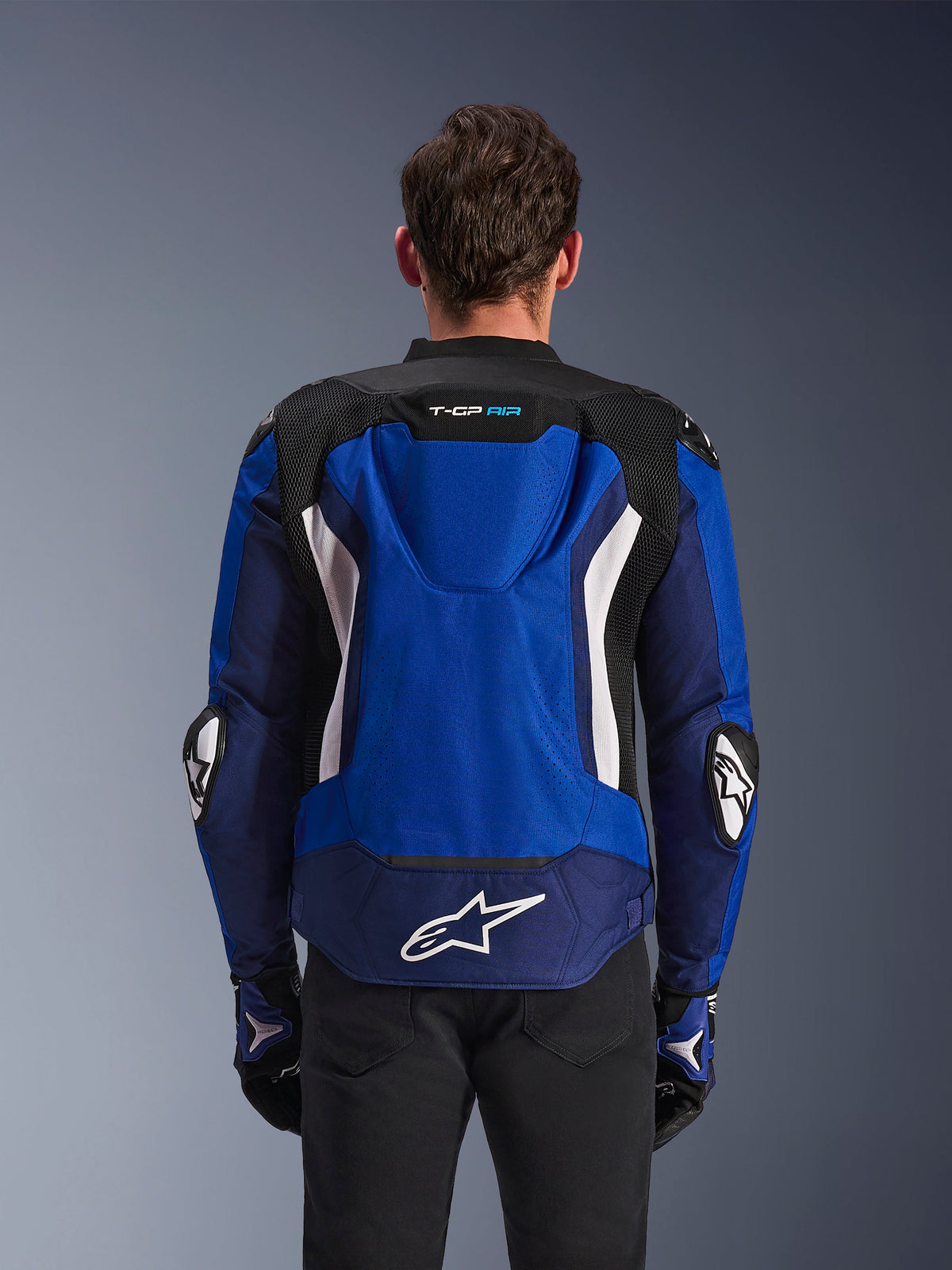 Alpinestars T-GP Air Jacke, Sport-Textil-Motorradjacke, Hellblau, Navy und Weiß, Rückansicht mit integriertem aerodynamischem Höcker und großem weißem Logo am unteren Rücken, getragen mit passenden blau-schwarzen Motorradhandschuhen